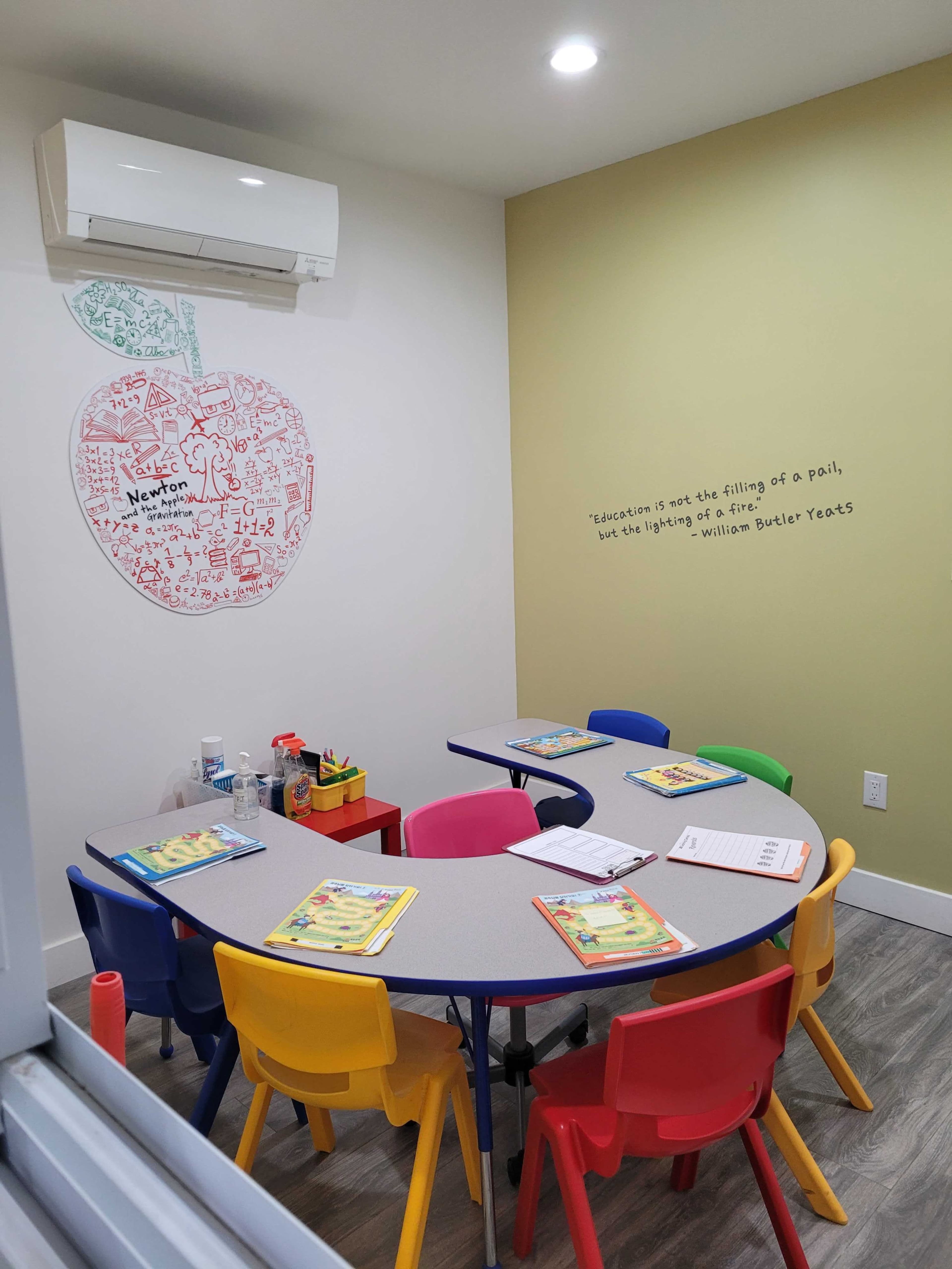 A colorful classroom features an apple-shaped mural, a round table surrounded by brightly colored chairs, and educational materials placed on the table.