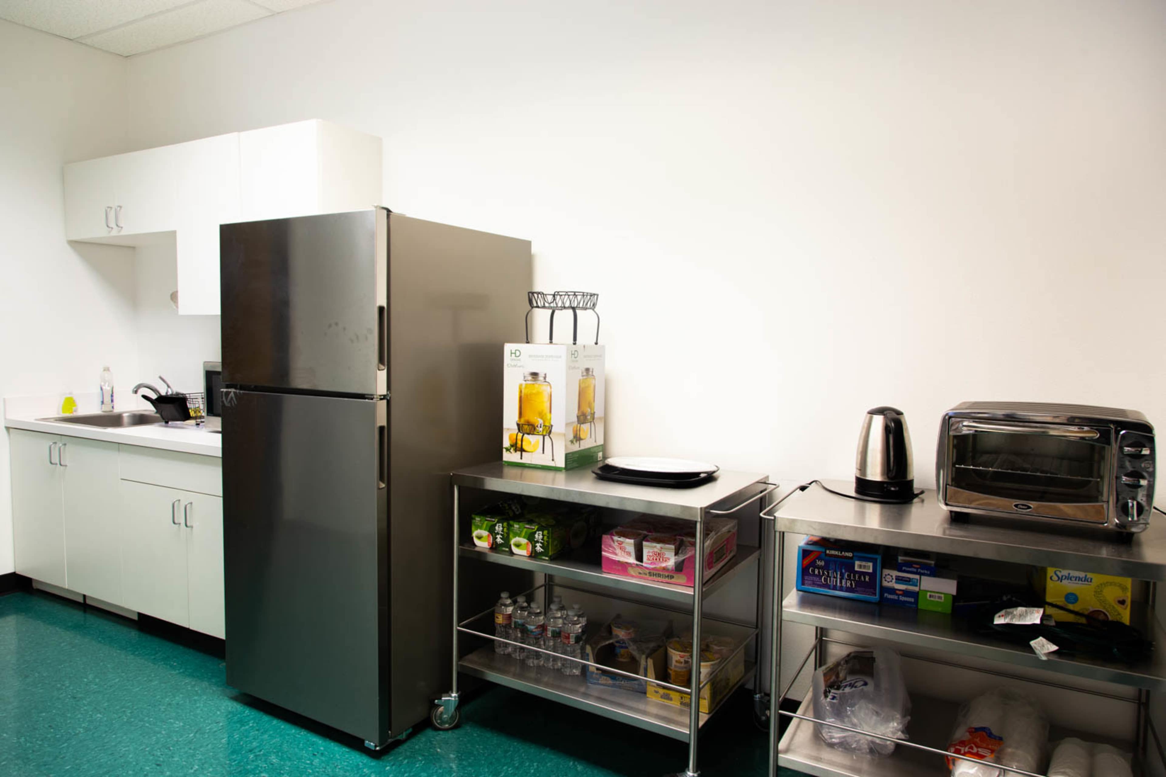A minimalist kitchen area features a stainless steel refrigerator, a microwave, and a shelving unit with various food and drink items.