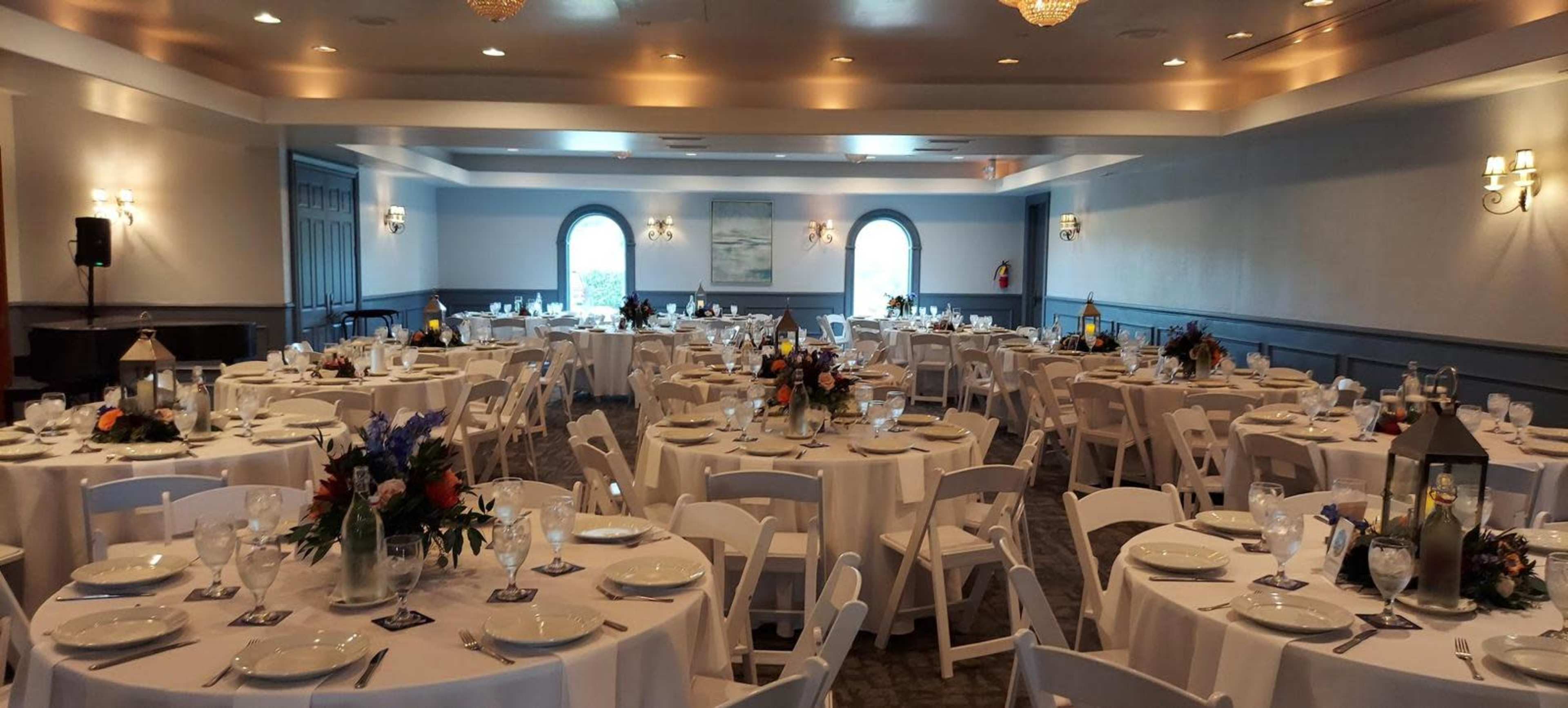 The image shows a banquet hall arranged for an event, with multiple round tables covered in white tablecloths and decorated with floral centerpieces.