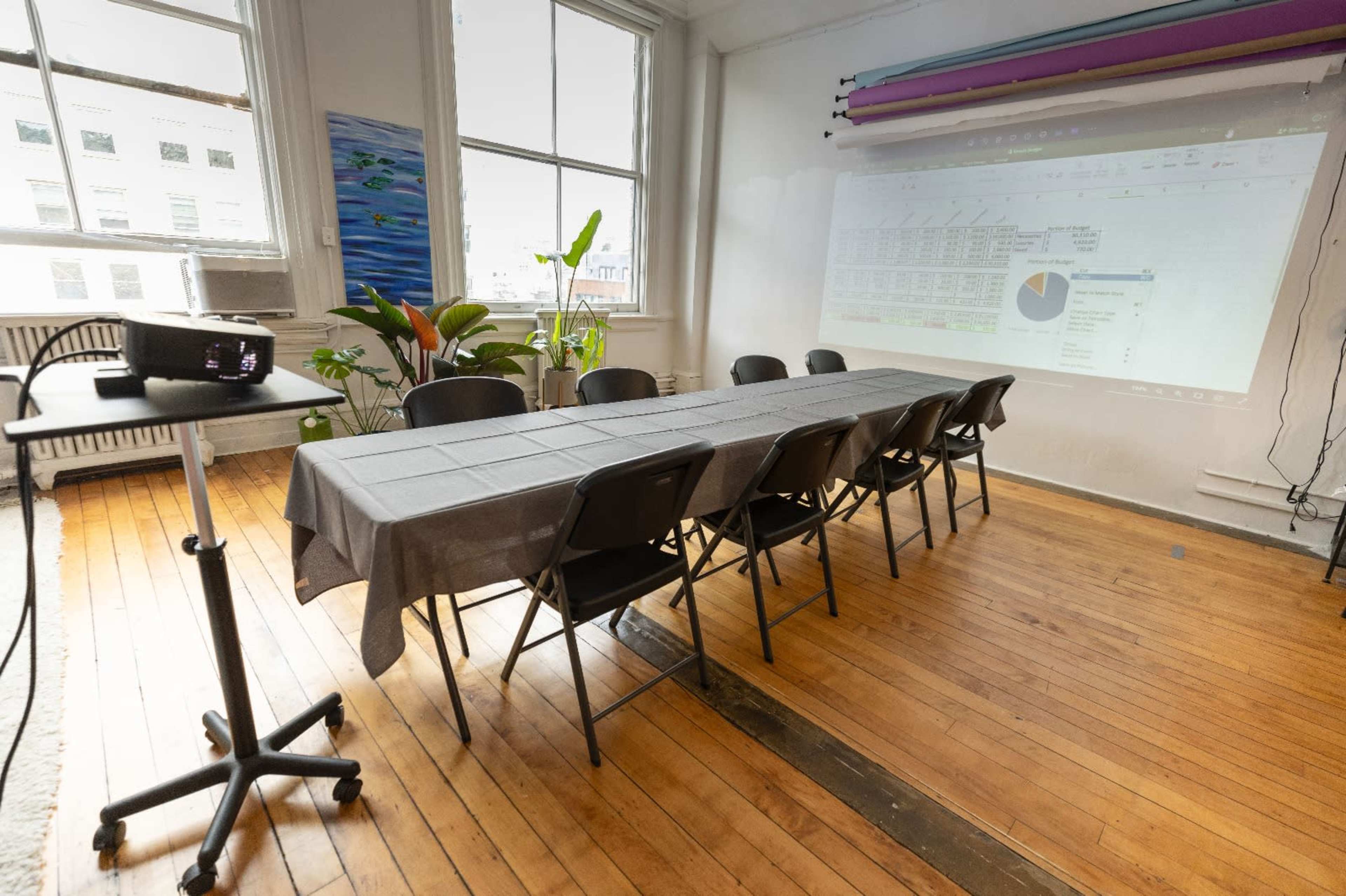 A long table with eight black chairs is set up in a well-lit room, featuring a projector displaying a chart on a wall.