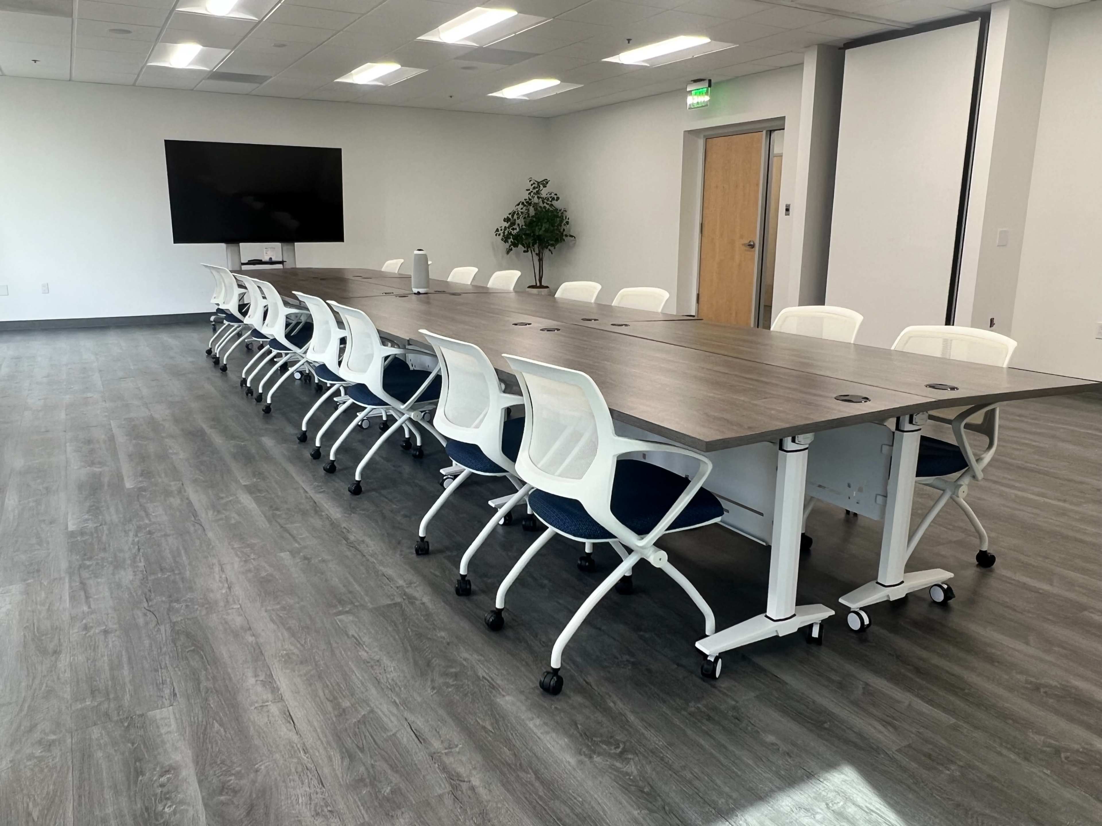 A long conference table with rolling white chairs is set up in a modern, brightly lit room that features a large screen and a potted plant.