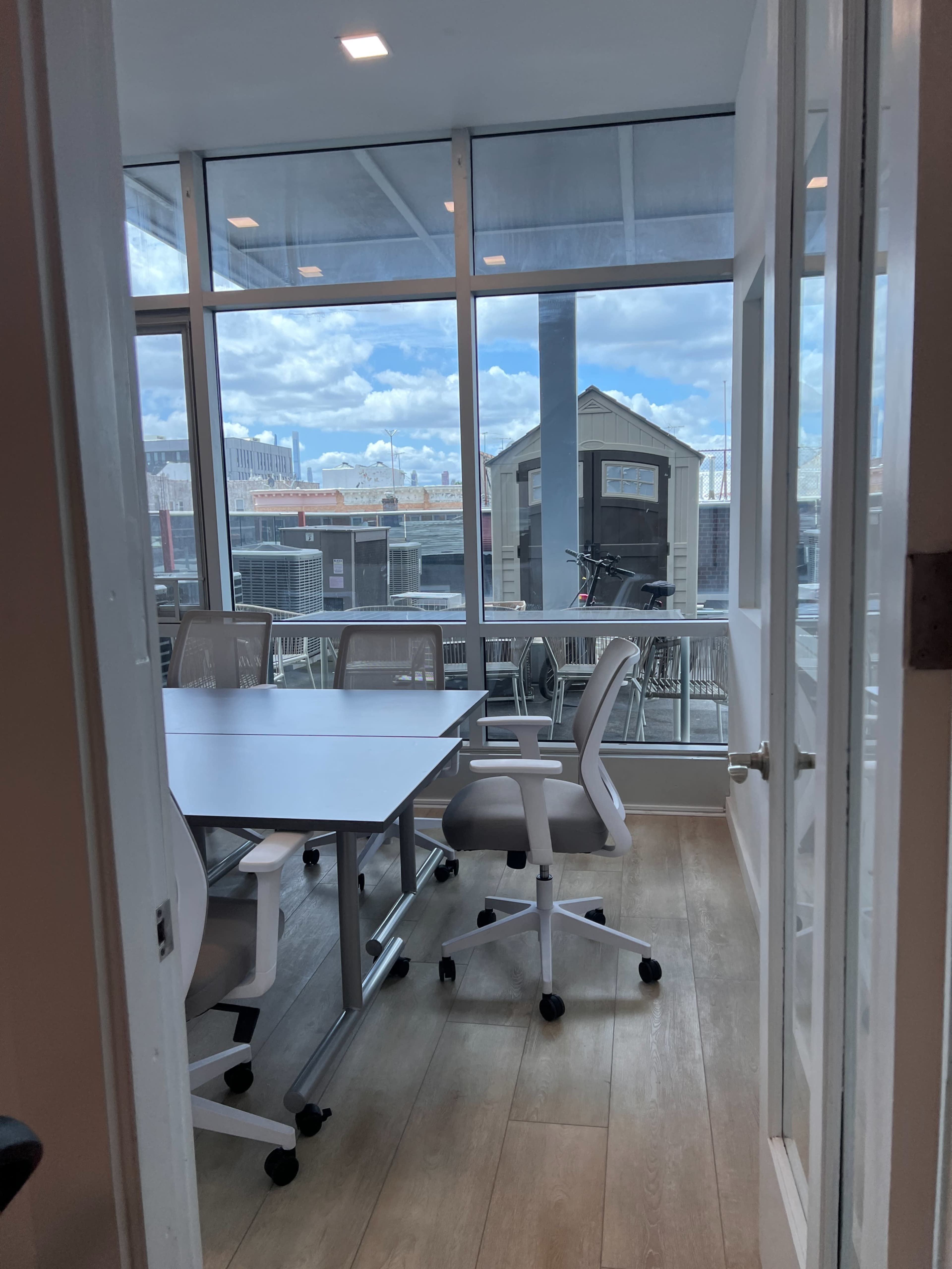A conference room with a white table and several chairs, viewed through a door, features large windows overlooking a cityscape.