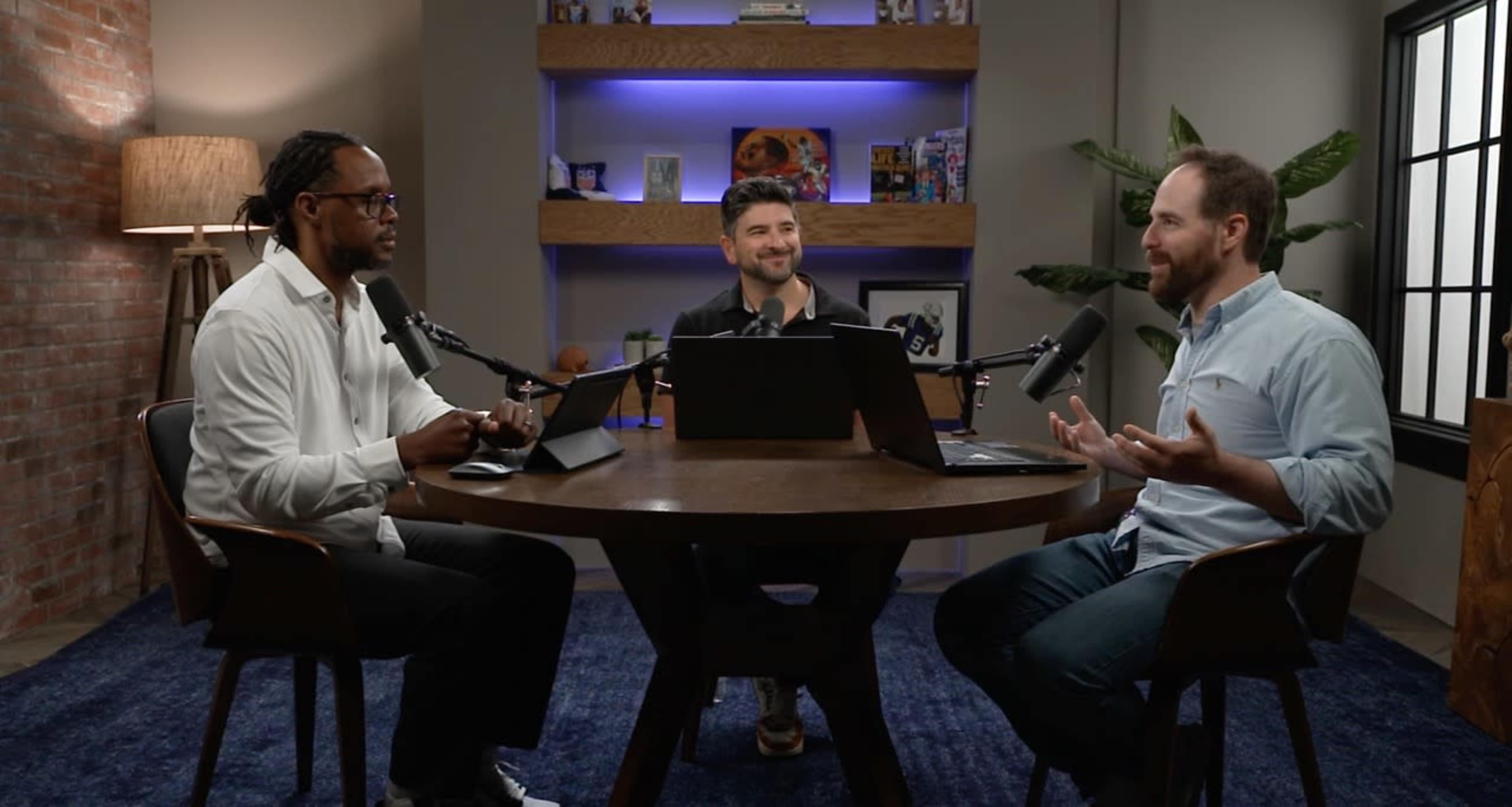 Three men are seated at a round table in a well-lit room, discussing while using laptops and microphones.