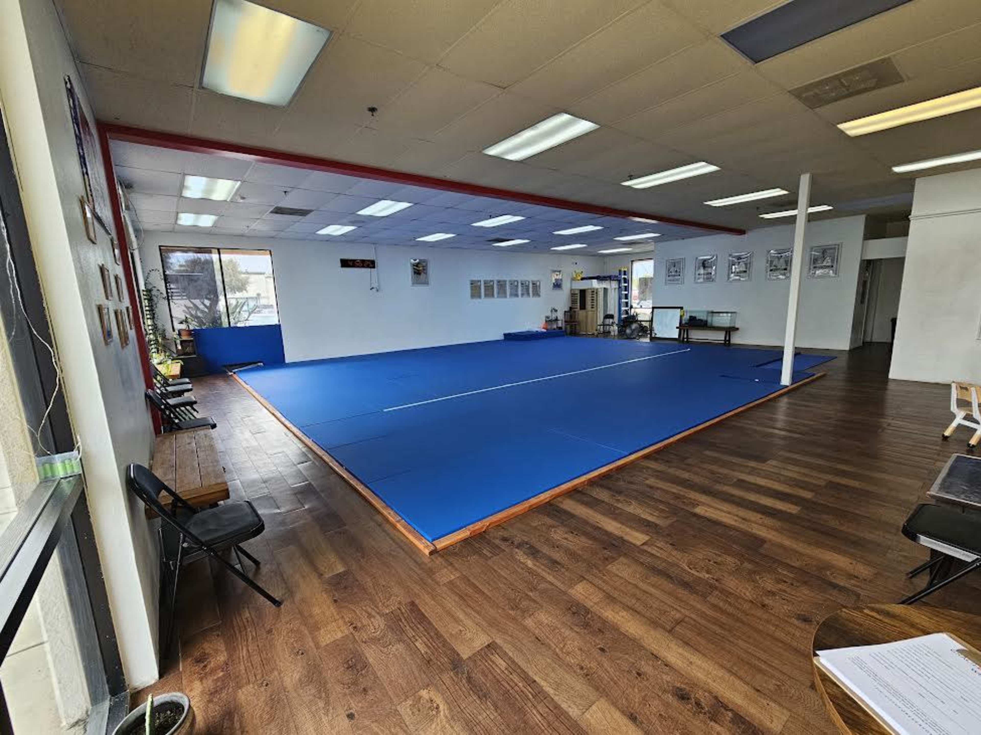 The image shows a spacious martial arts training area with blue mats, wooden flooring, and a few chairs along the walls.