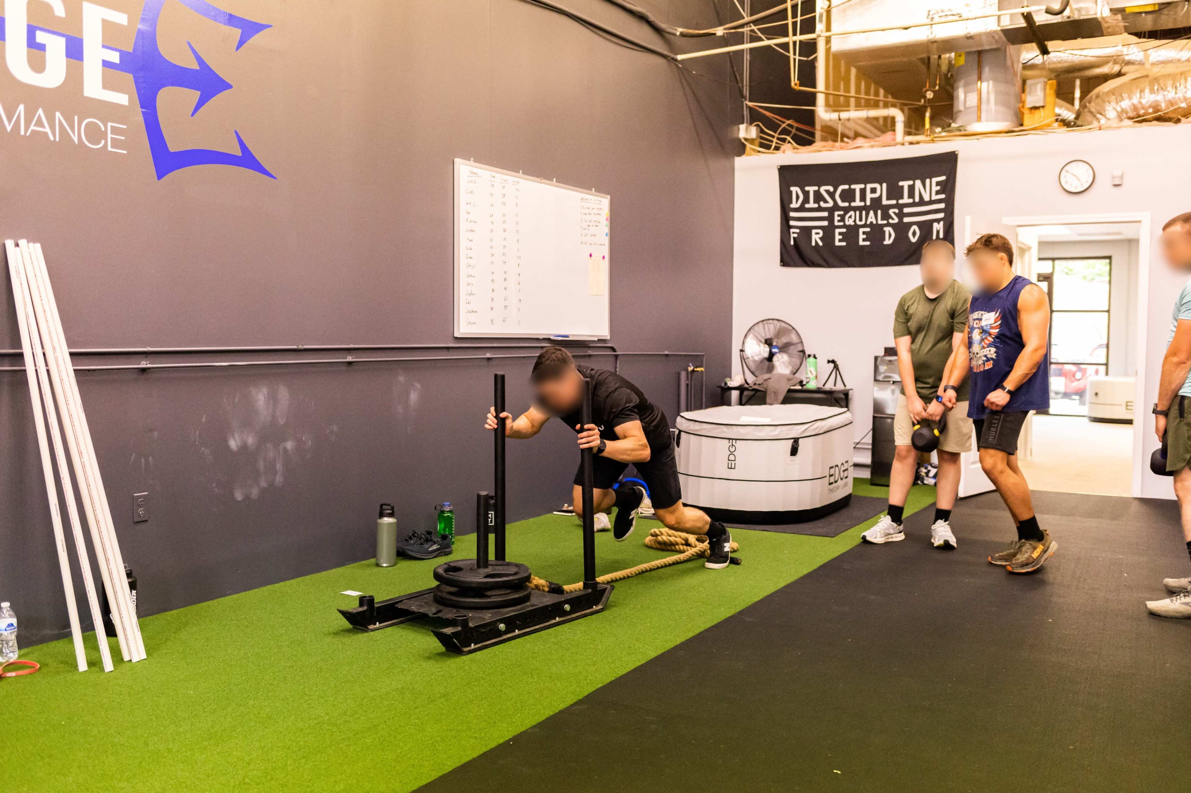 A person is pushing a weighted sled across a gym floor while others observe and participate in the workout.