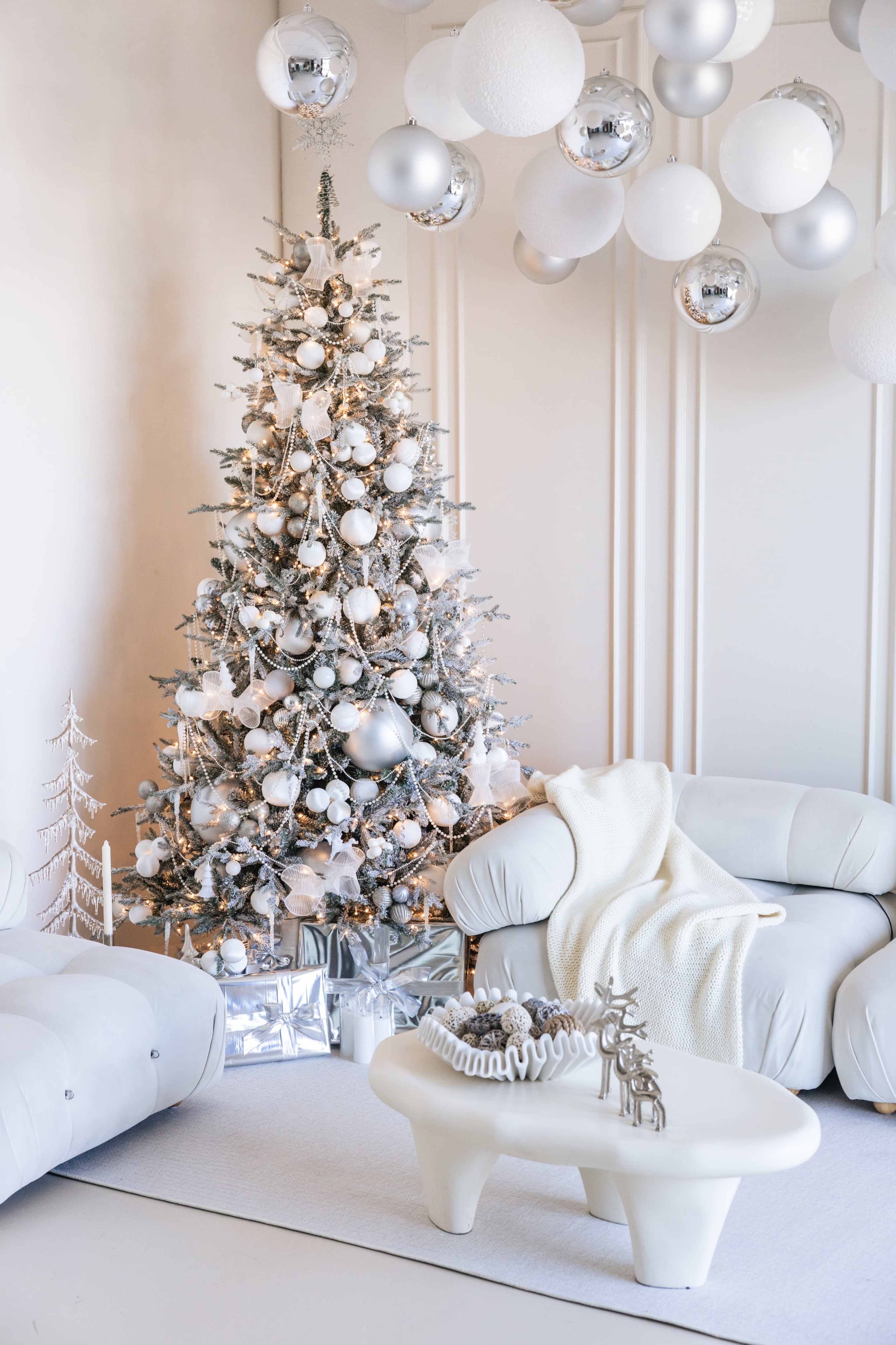 A decorated Christmas tree with silver ornaments and lights is placed in a stylish living room with modern furniture and white decorations.