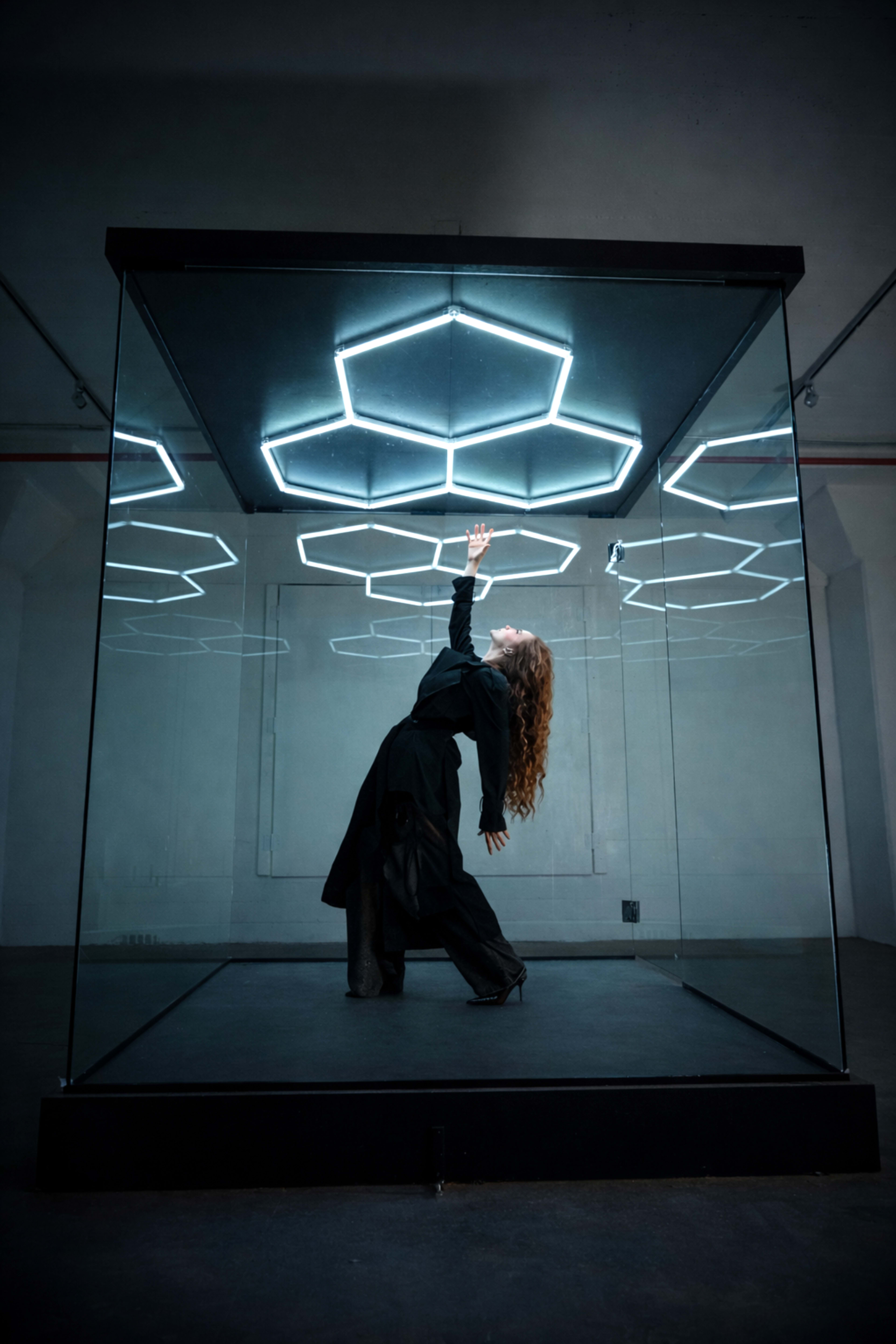 A person with long, curly hair poses dramatically inside a glass cube illuminated by hexagonal LED lights on the ceiling.
