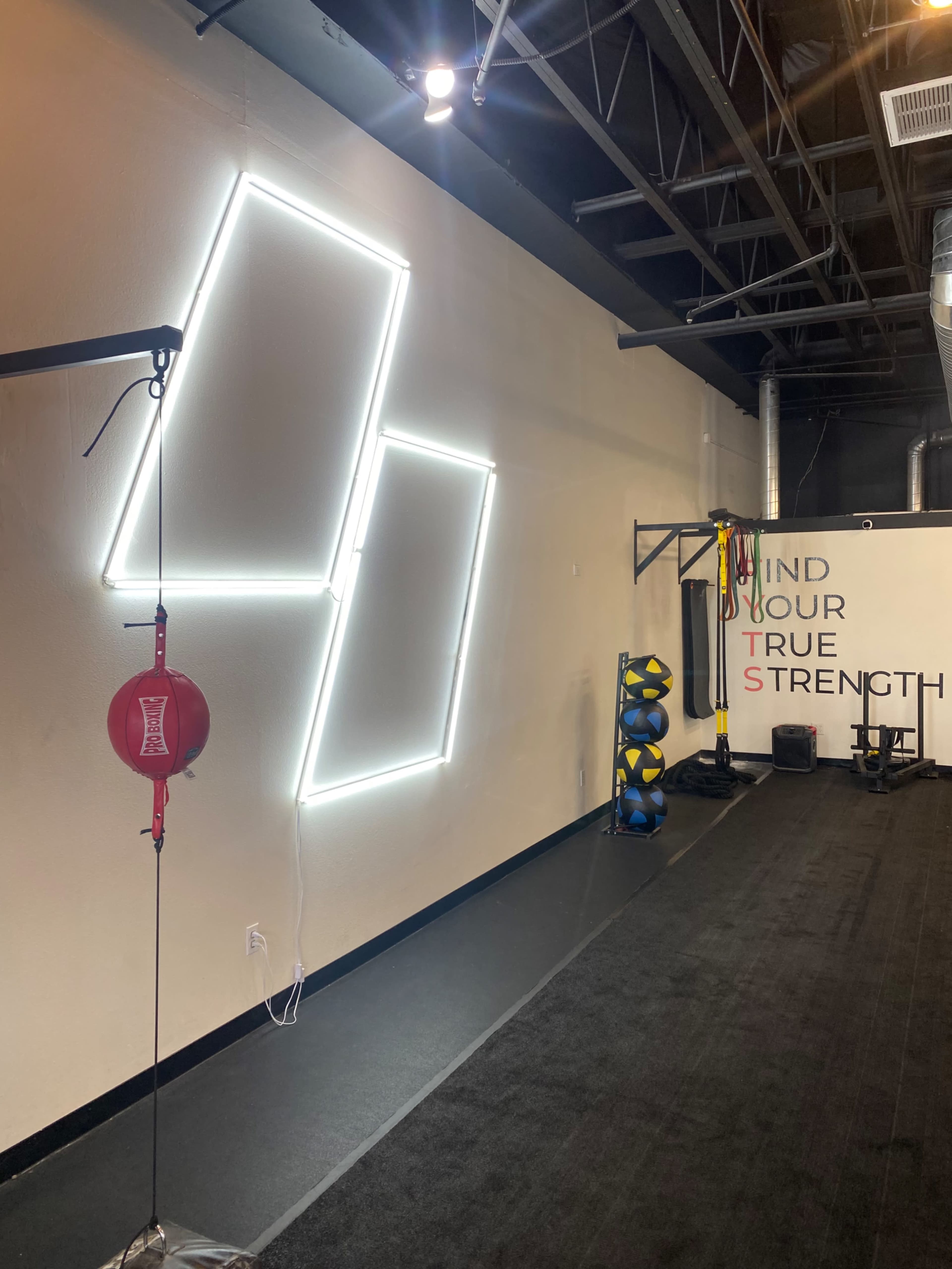 A fitness studio with a minimalist design, featuring a wall with three illuminated rectangular shapes, a boxing bag hanging from the ceiling, and fitness equipment arranged on the floor.