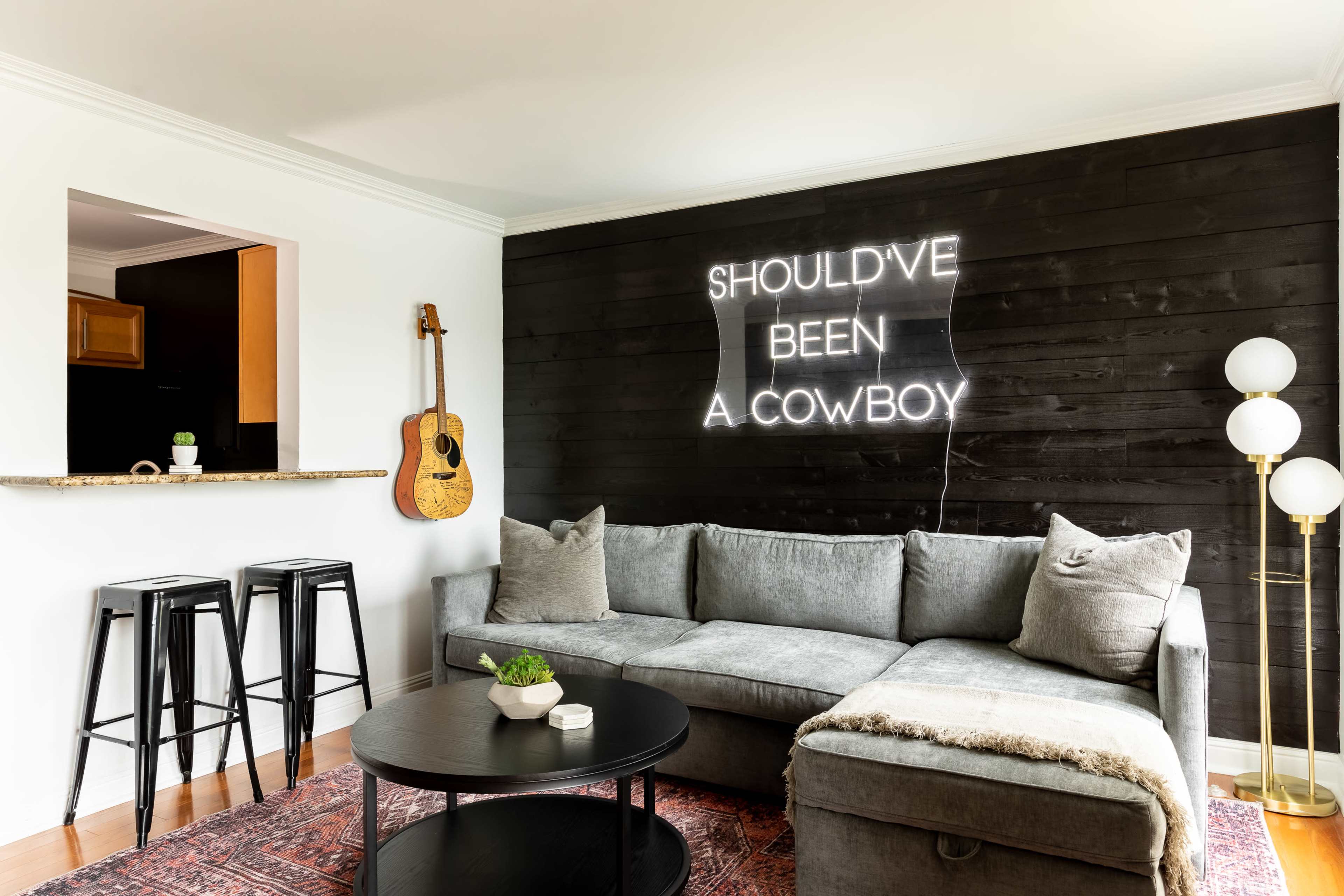 A cozy living room features a gray sectional sofa, a black circular table, and a neon sign on the wall that reads "SHOULDN'T HAVE BEEN A COWBOY."