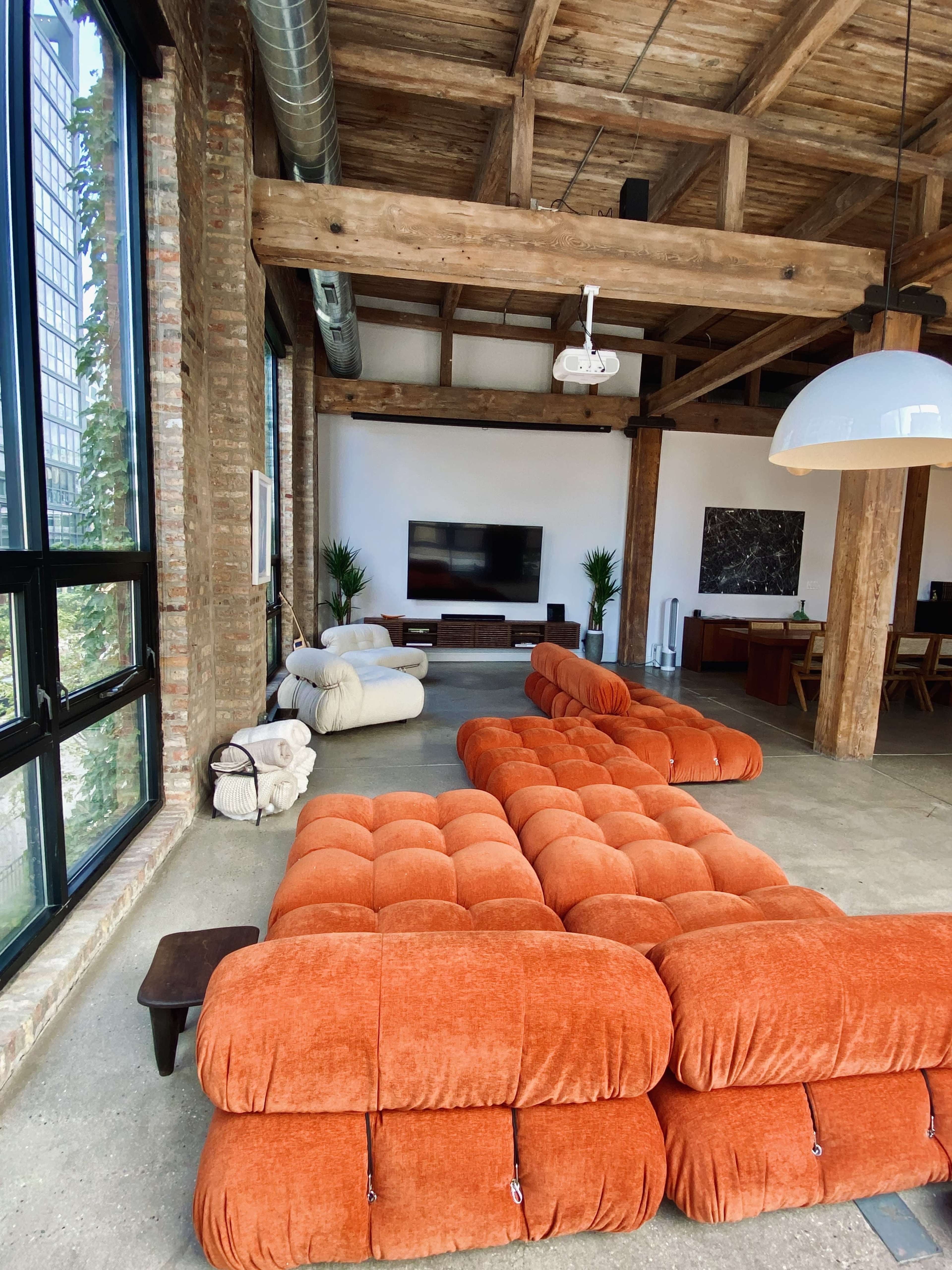 A spacious, modern living area featuring large windows, exposed wooden beams, and orange tufted sofas arranged along a concrete floor.