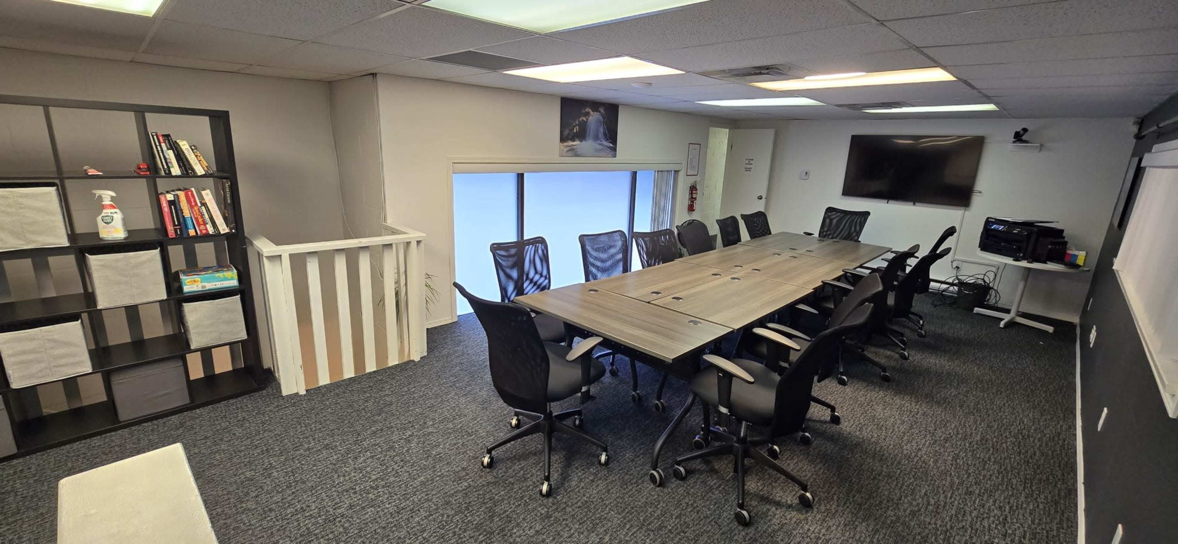 A spacious conference room featuring a long table surrounded by black office chairs, a bookshelf on the left, and a television mounted on the wall.