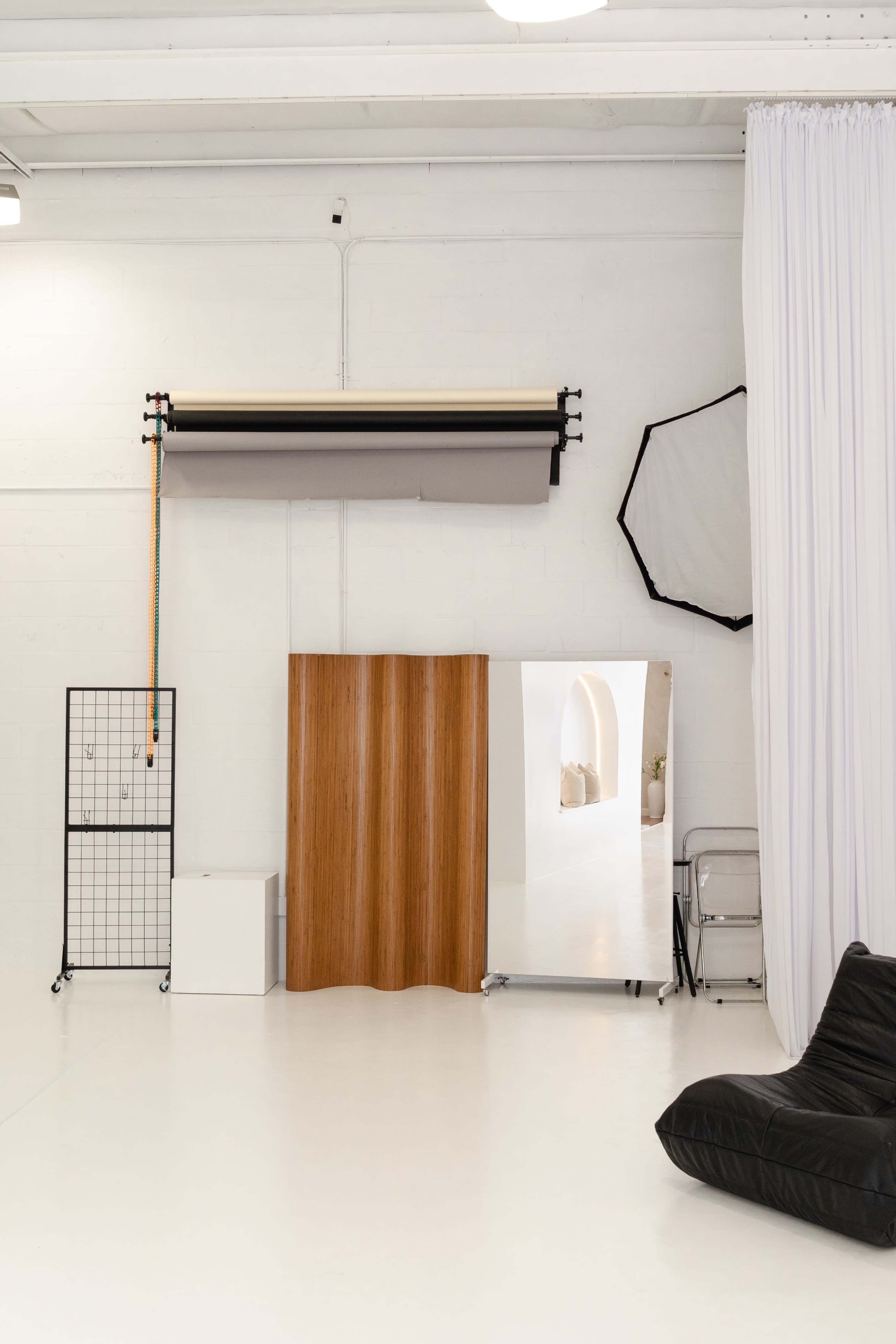 The image showcases a well-lit photography studio featuring a wooden backdrop, a large mirror, a fabric drape, and a black lounge chair.