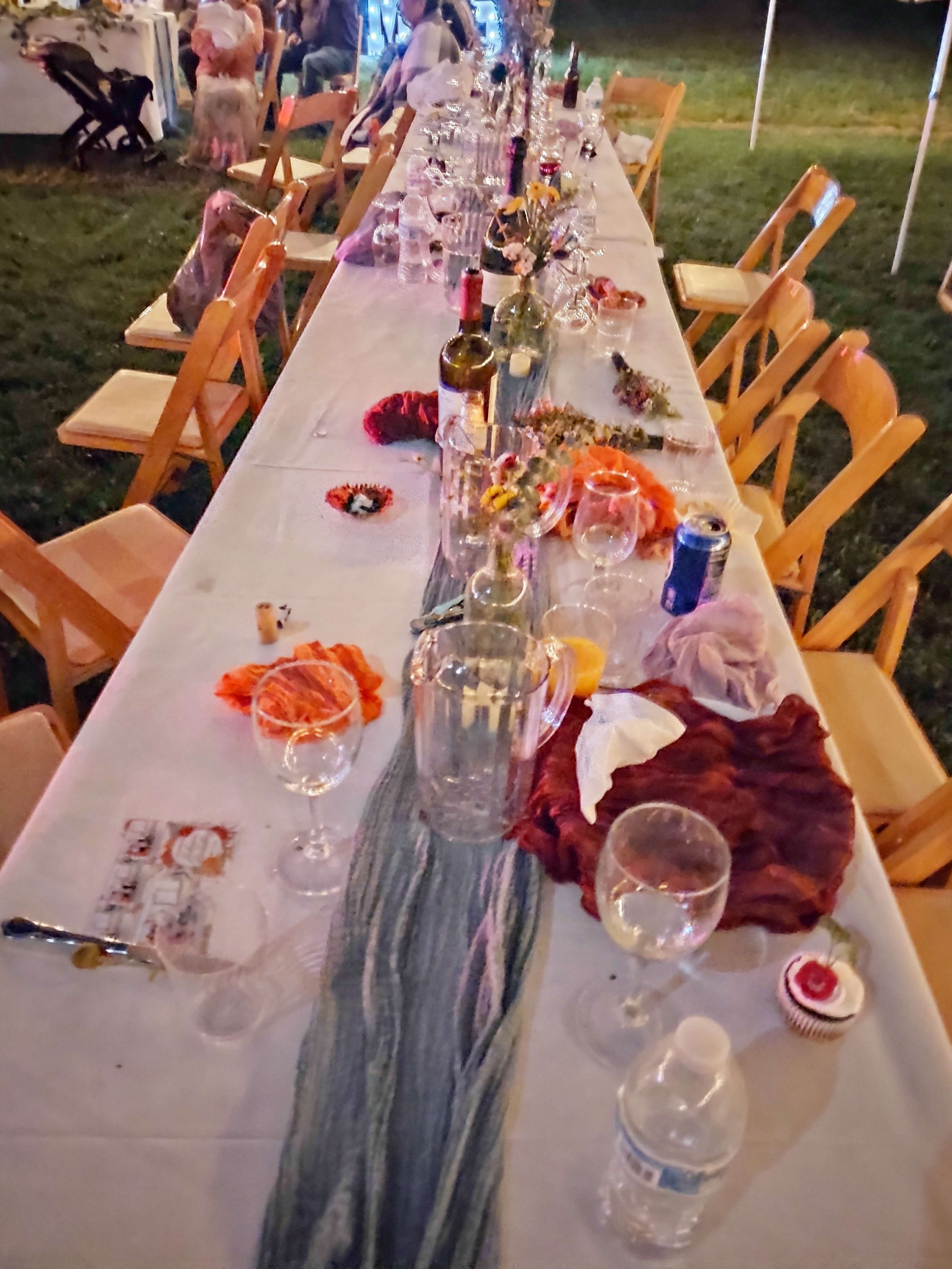 A long table is set outdoors, covered with empty glasses, wine bottles, and scattered decorations, surrounded by wooden chairs.