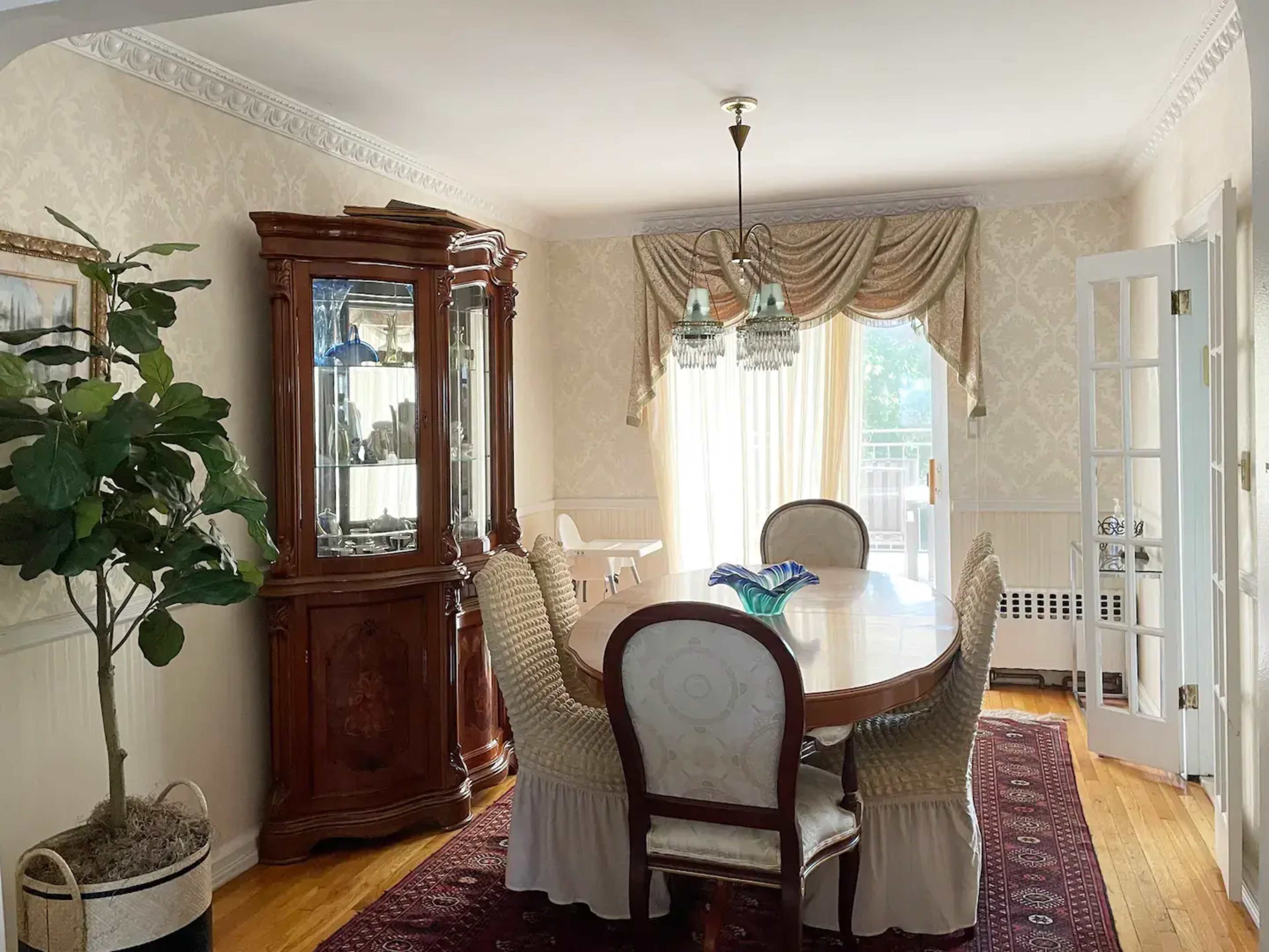 A dining room featuring a large, round table surrounded by upholstered chairs, a china cabinet, and natural light streaming through the draped windows.