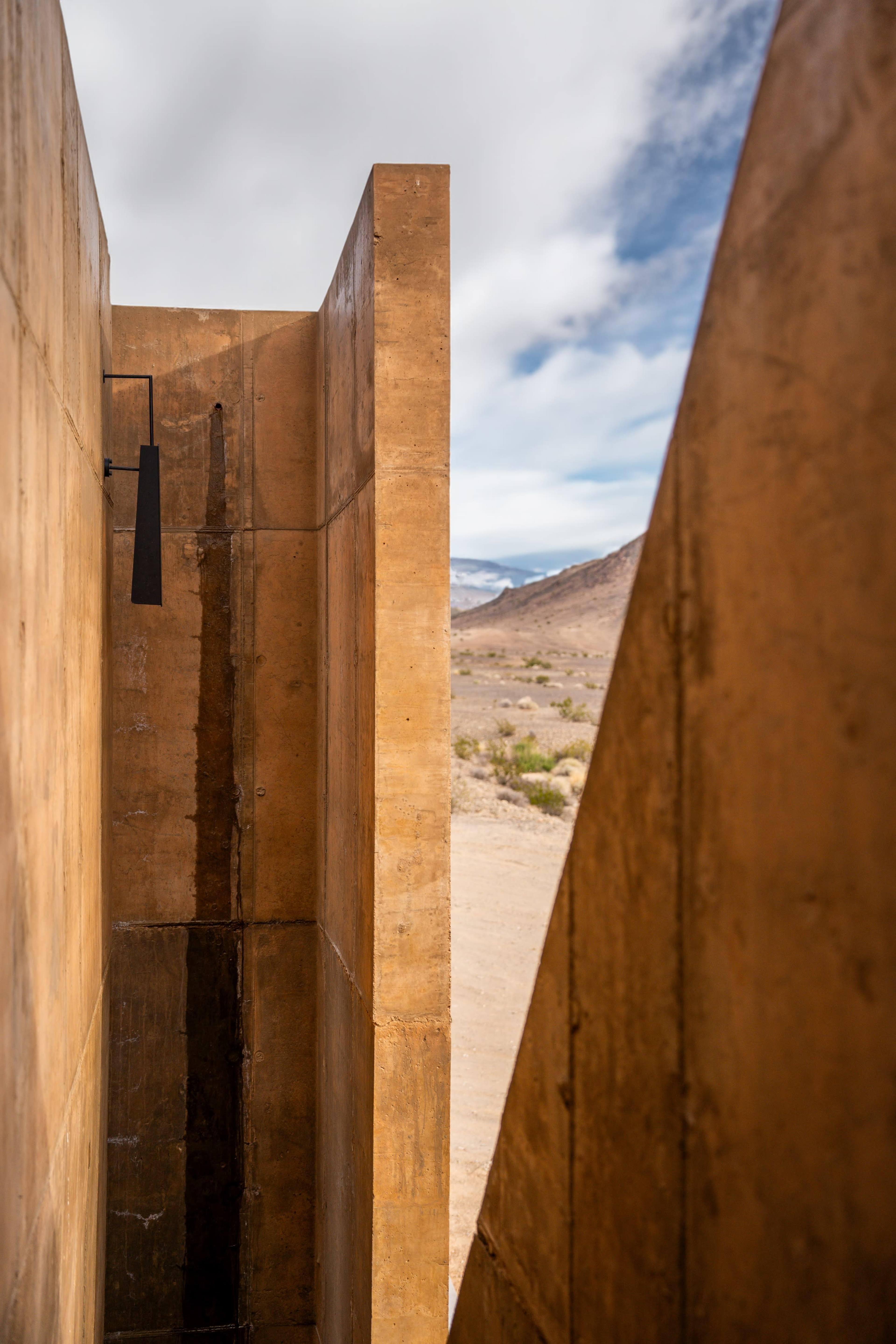 A narrow corridor framed by large, concrete walls, leading towards a view of the desert landscape under a cloudy sky.