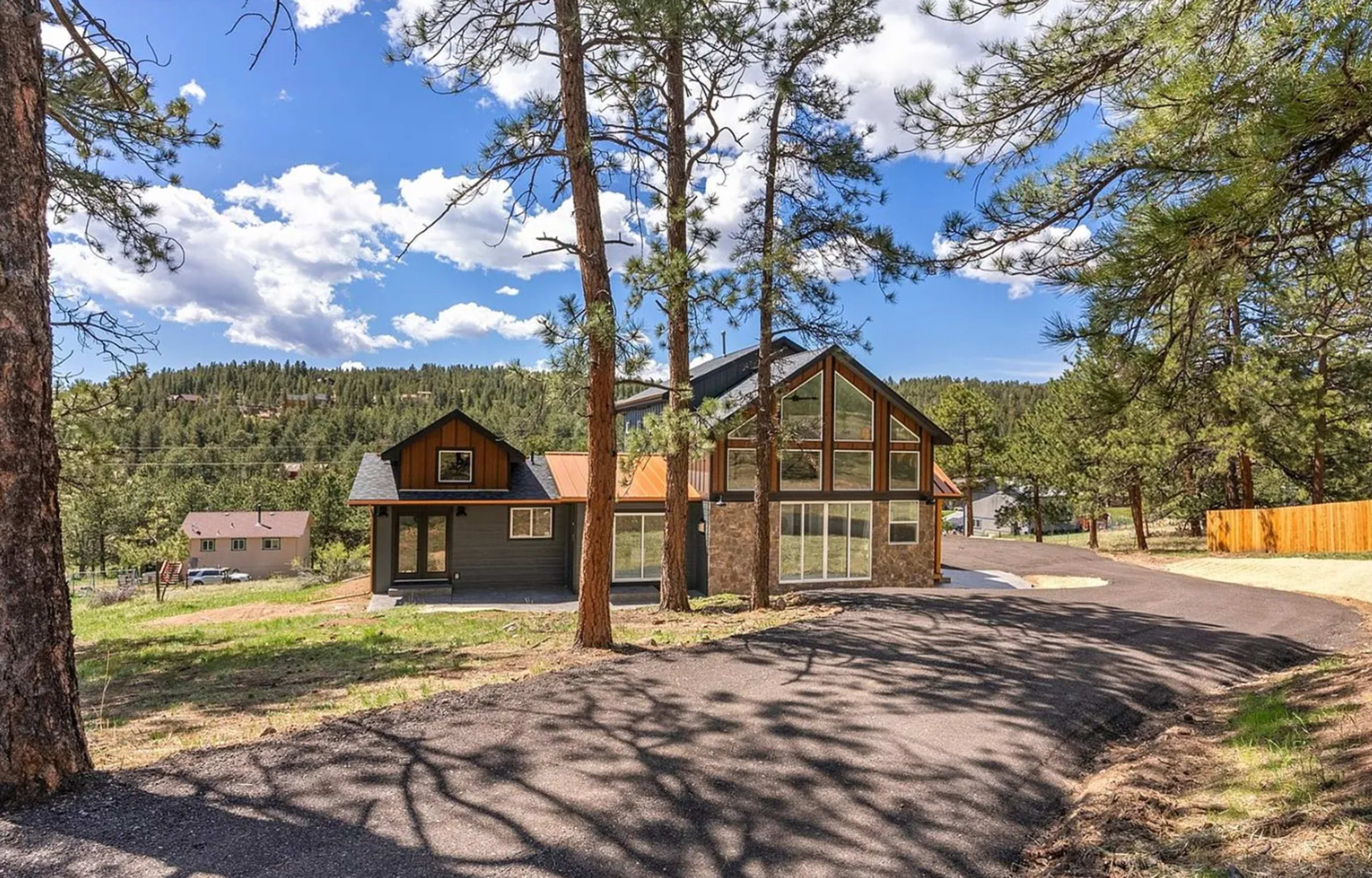 A two-story house with large windows and a stone facade is set among tall pine trees on a paved driveway.