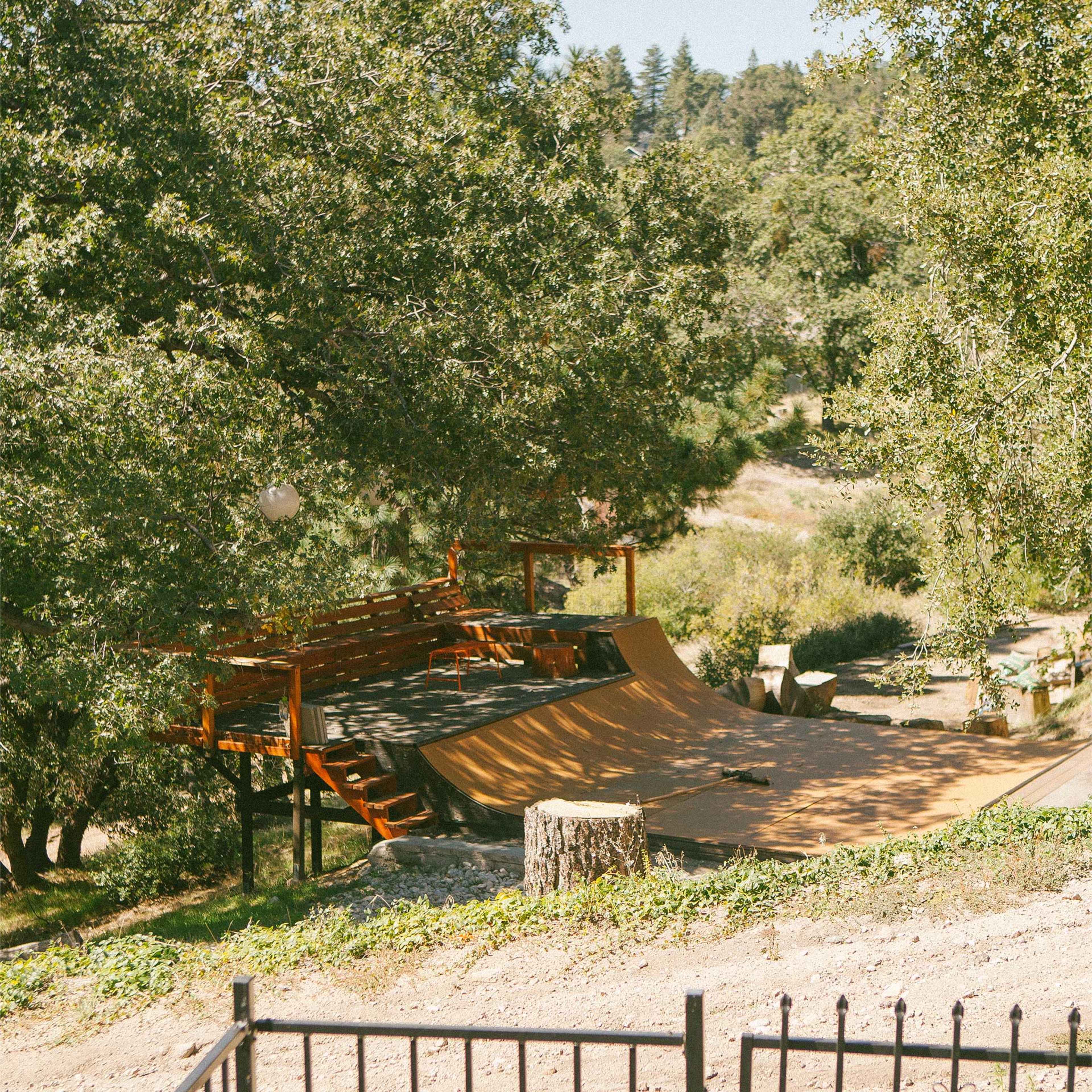 A wooden skate ramp is situated in a natural outdoor setting surrounded by trees and a gravel path.