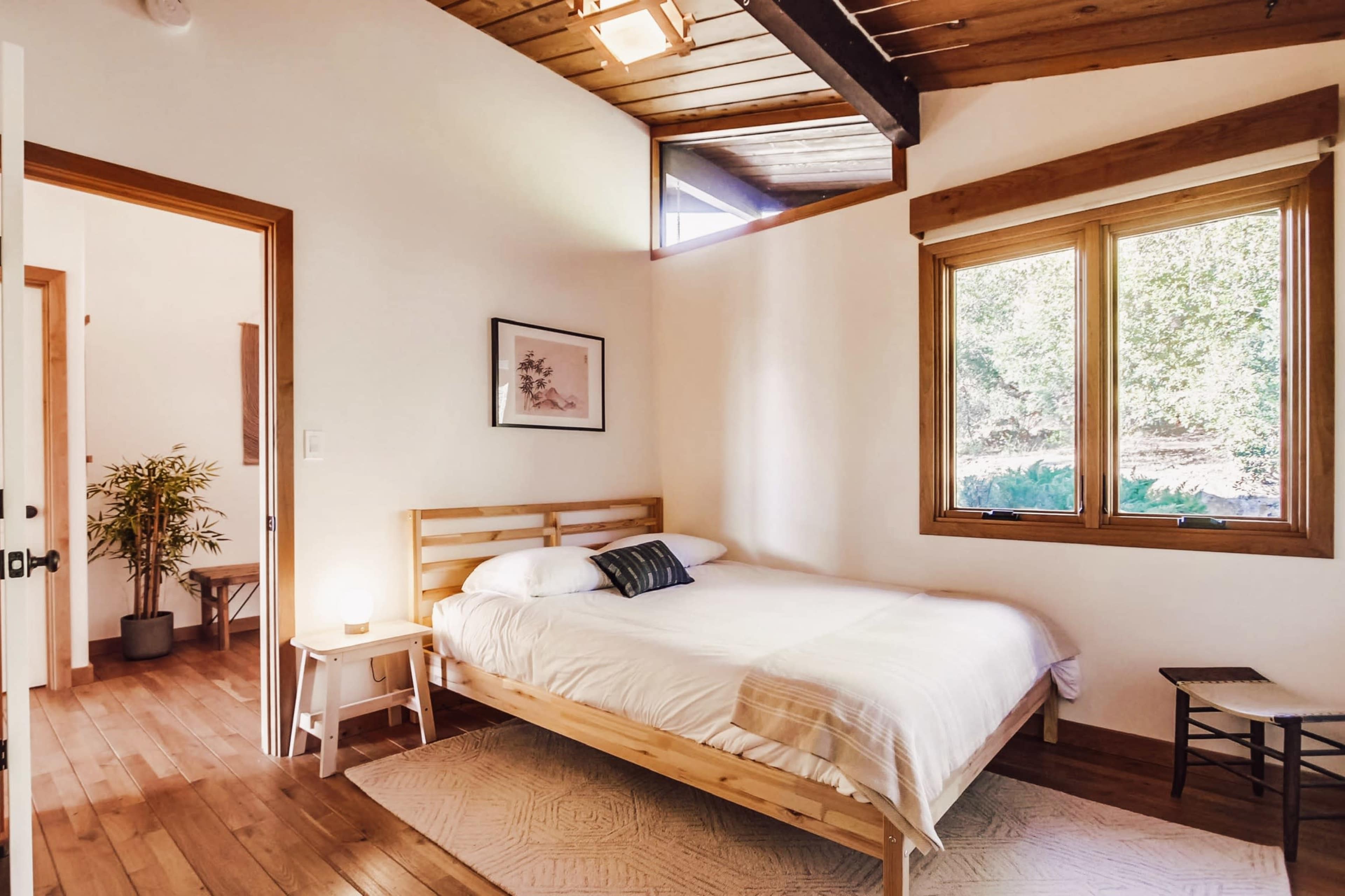 A simple bedroom features a wooden bed with white bedding, a small bedside lamp, and windows that provide natural light.