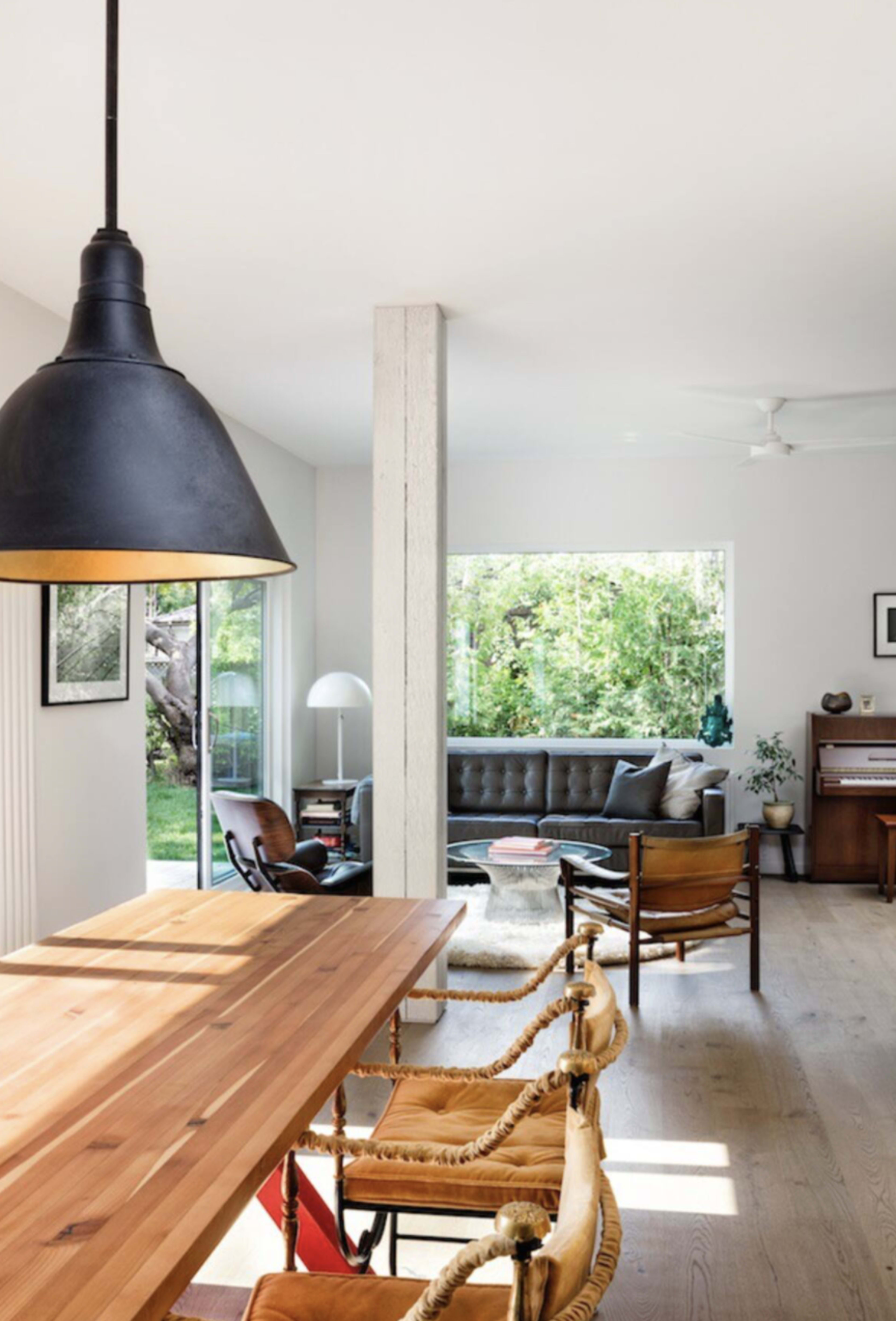 A wooden dining table with stylish chairs is positioned in a bright, open-plan living space featuring a large window that overlooks a verdant garden.