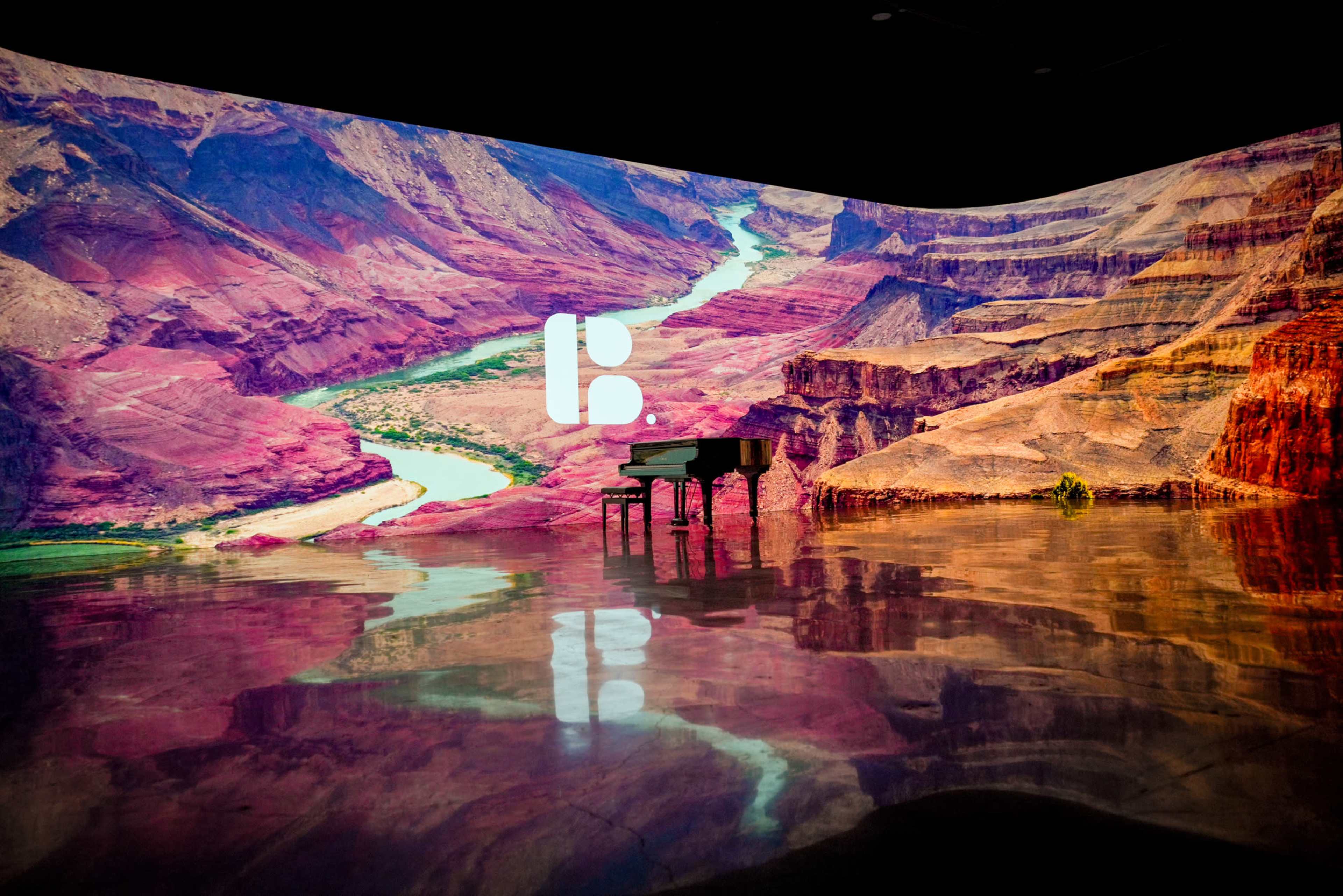 A grand piano stands in front of a large, colorful projection of a canyon landscape with a river winding through it.