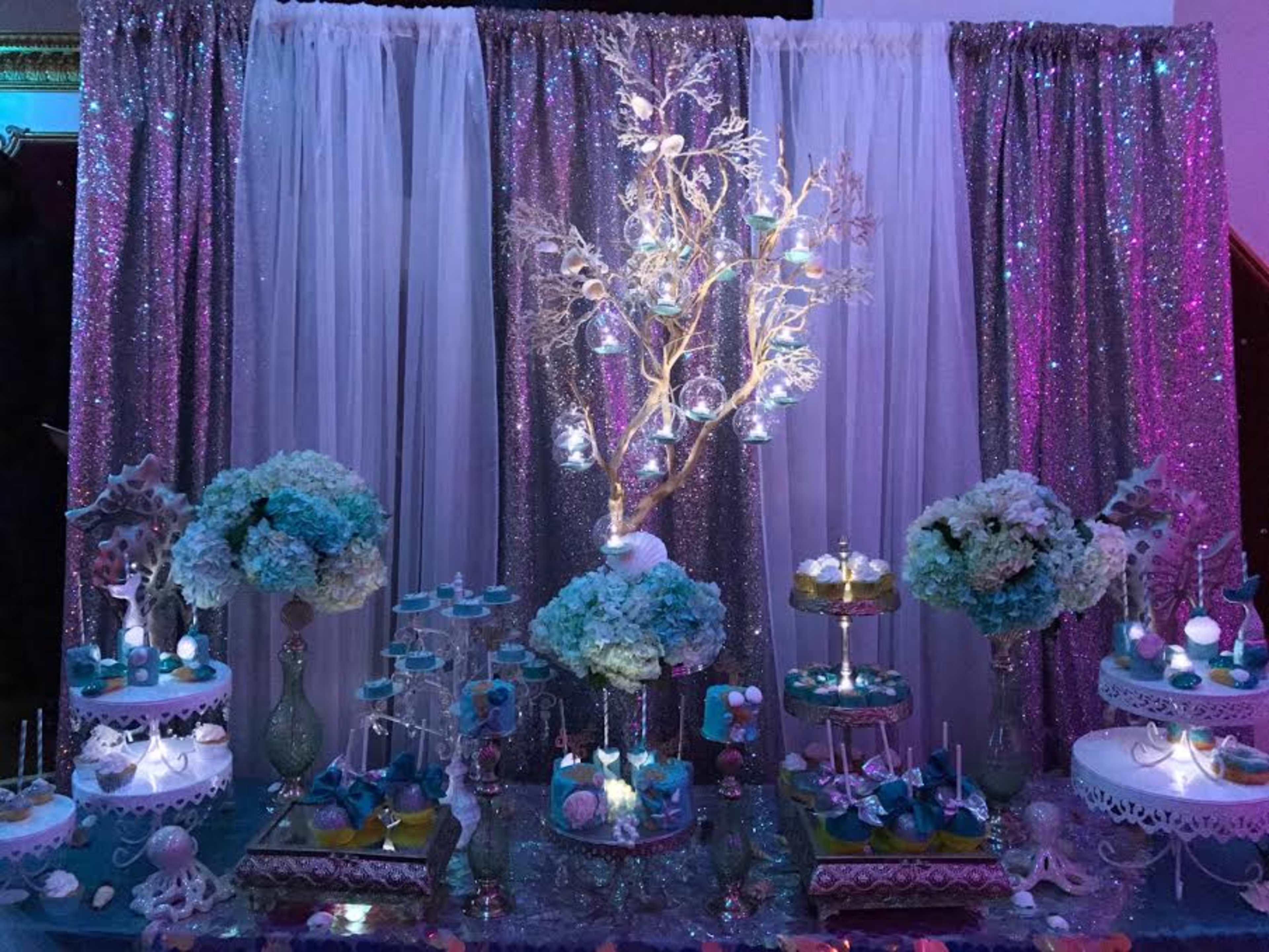 A decorated dessert table featuring tiered trays of colorful treats, floral arrangements, and illuminated decorative elements against a backdrop of sparkling fabrics.