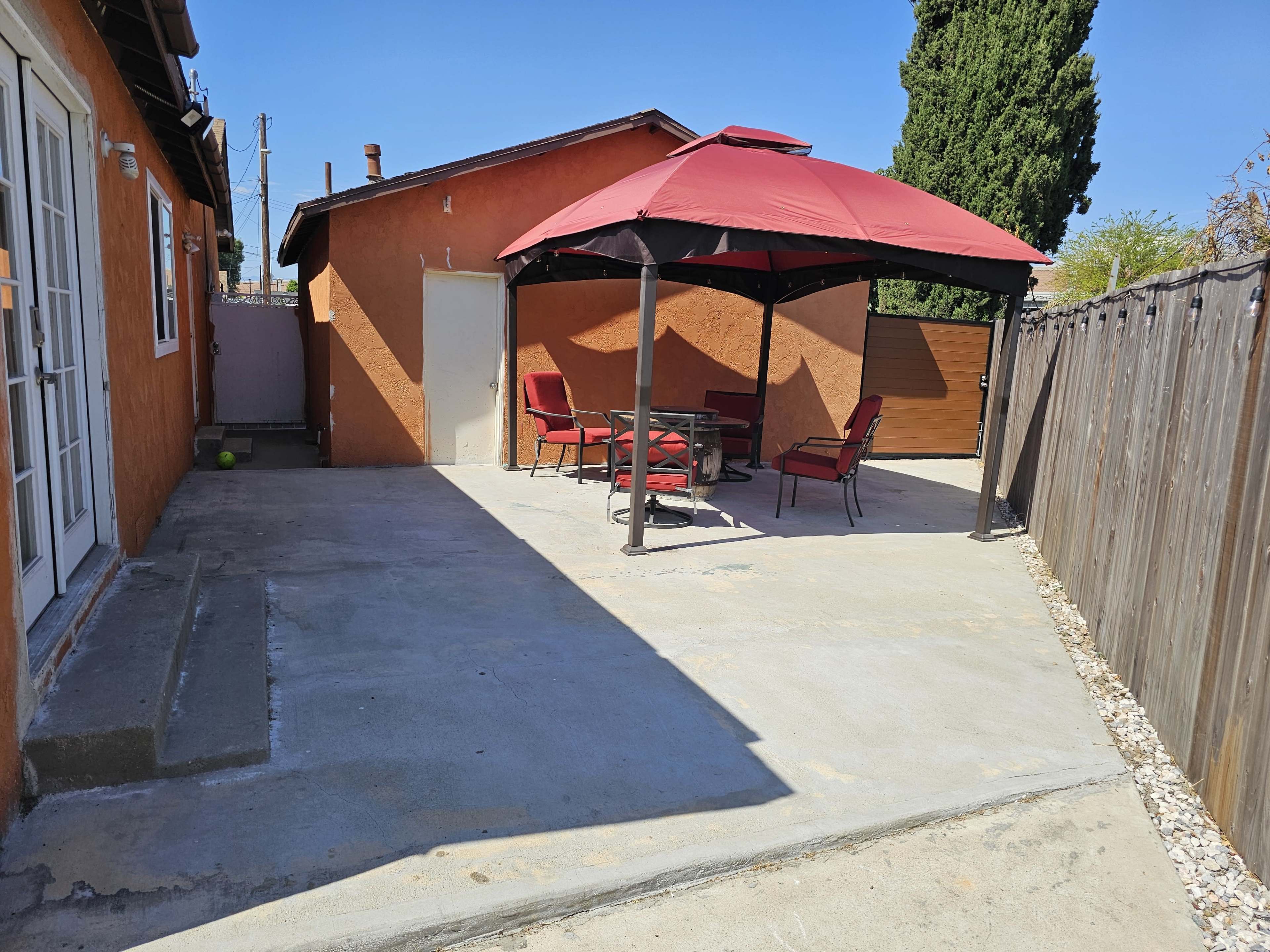 A patio area with a red gazebo and four chairs set around a table, adjacent to an orange-colored building and surrounded by a wooden fence.