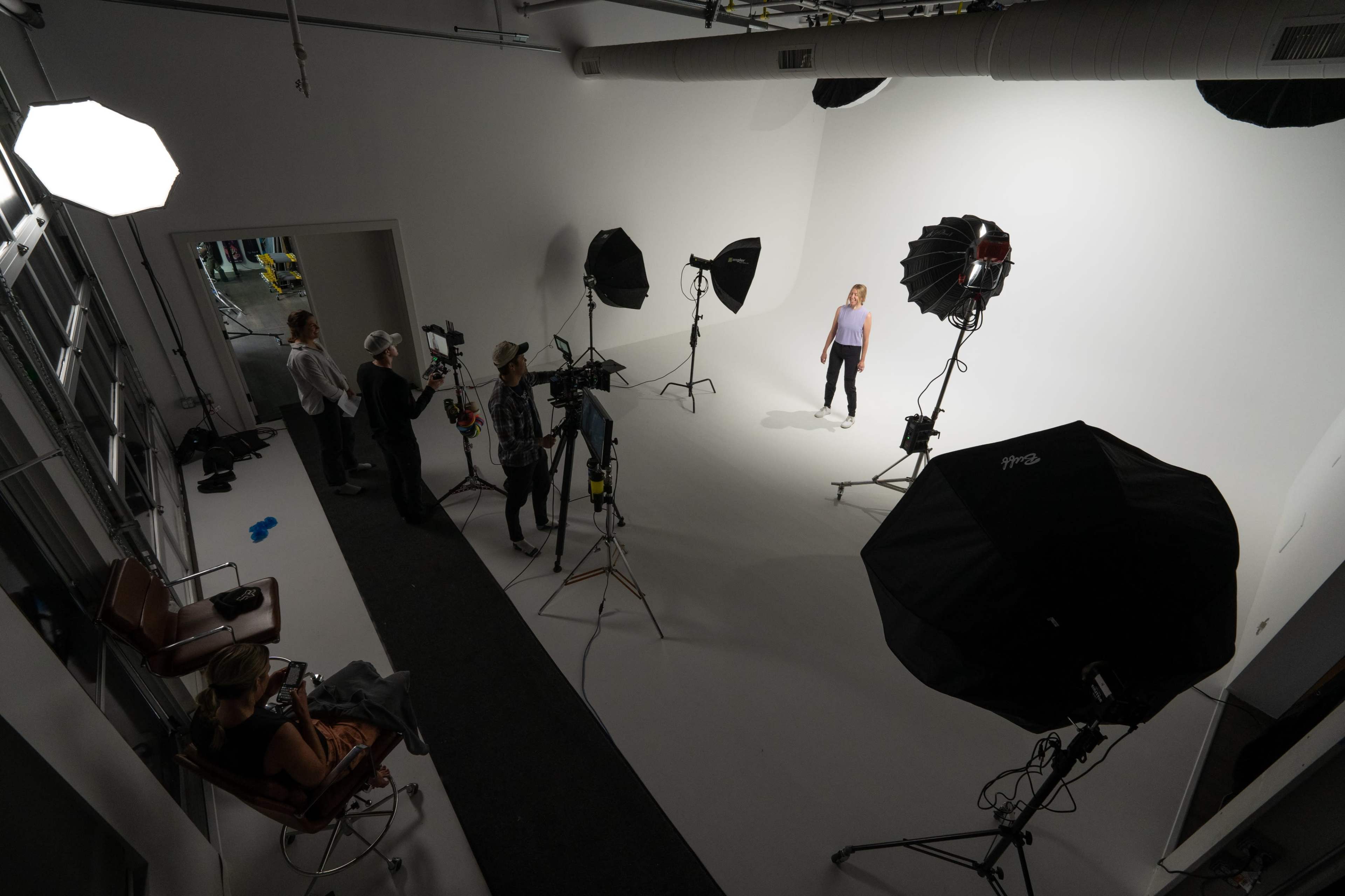 A photography studio with multiple lights and a model posing in front of a blank white backdrop, while a crew sets up equipment nearby.