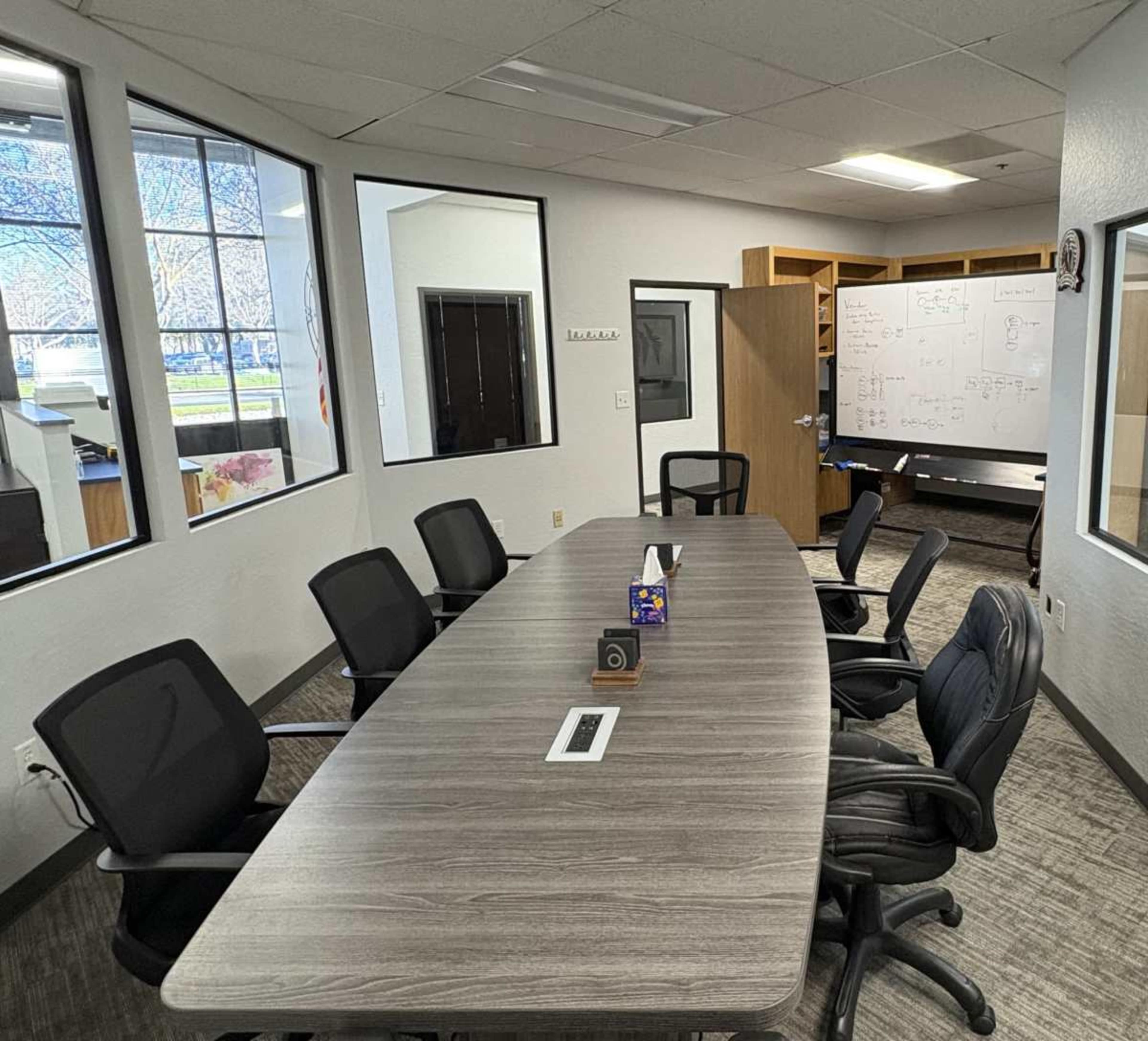 The image shows a conference room with a long table surrounded by several black chairs, large windows letting in natural light, and a whiteboard visible in the background.