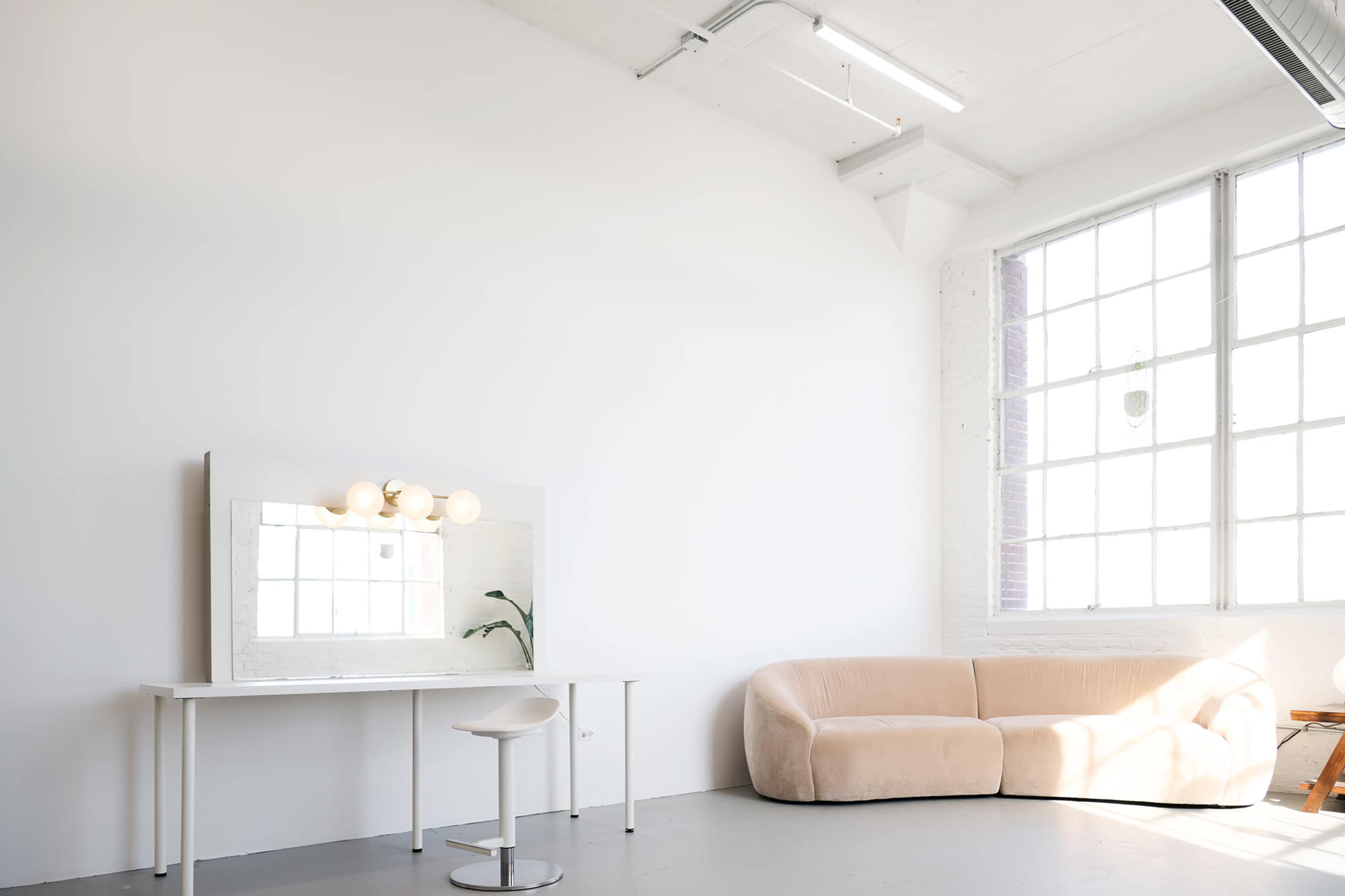 A bright, minimalist room featuring a light-colored sofa, a simple table with a mirror and round bulbs, and large windows allowing natural light.