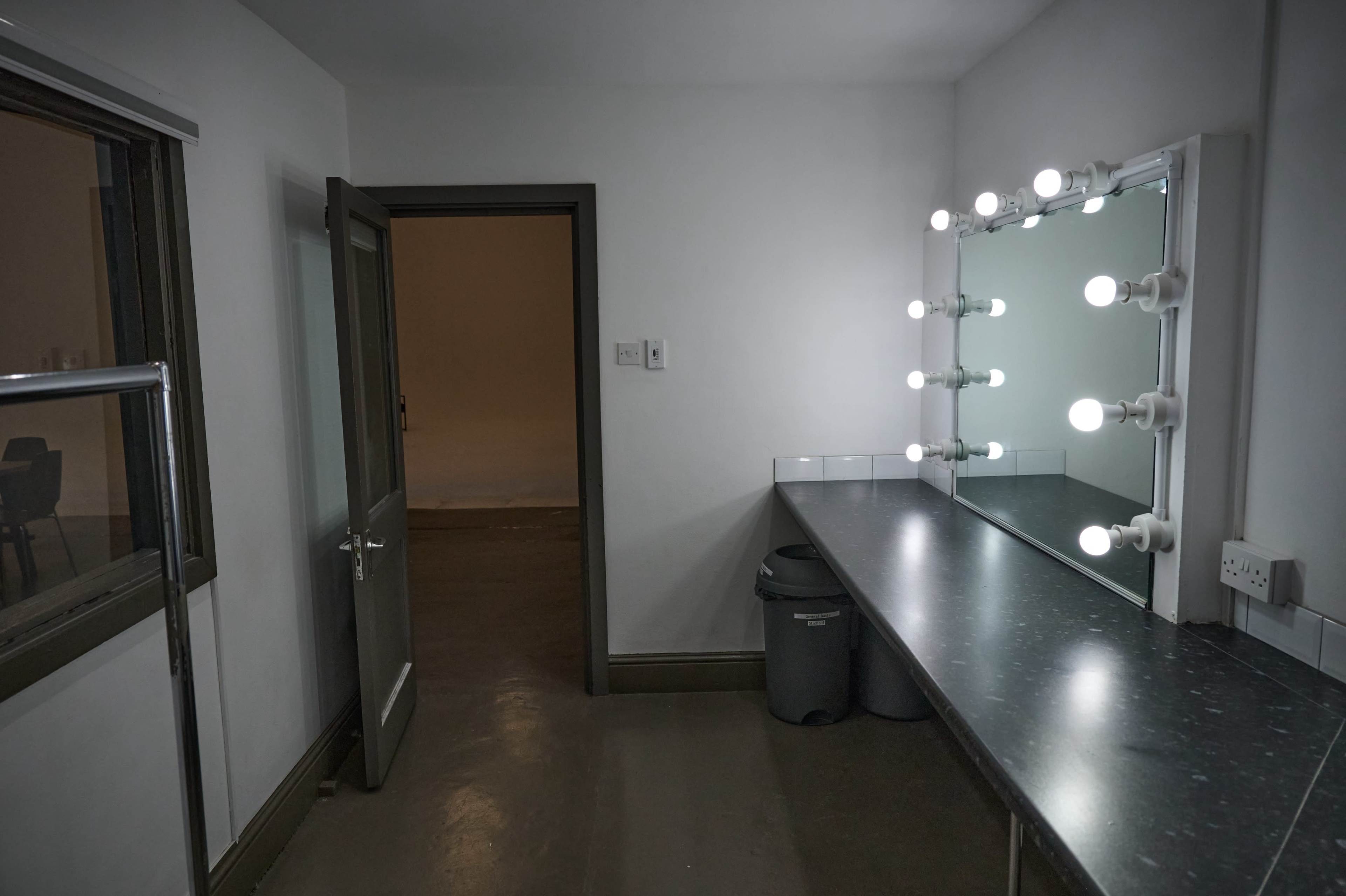 The image shows a well-lit dressing room featuring a large mirror with adjustable lights, a countertop, and a gray door that leads outside.