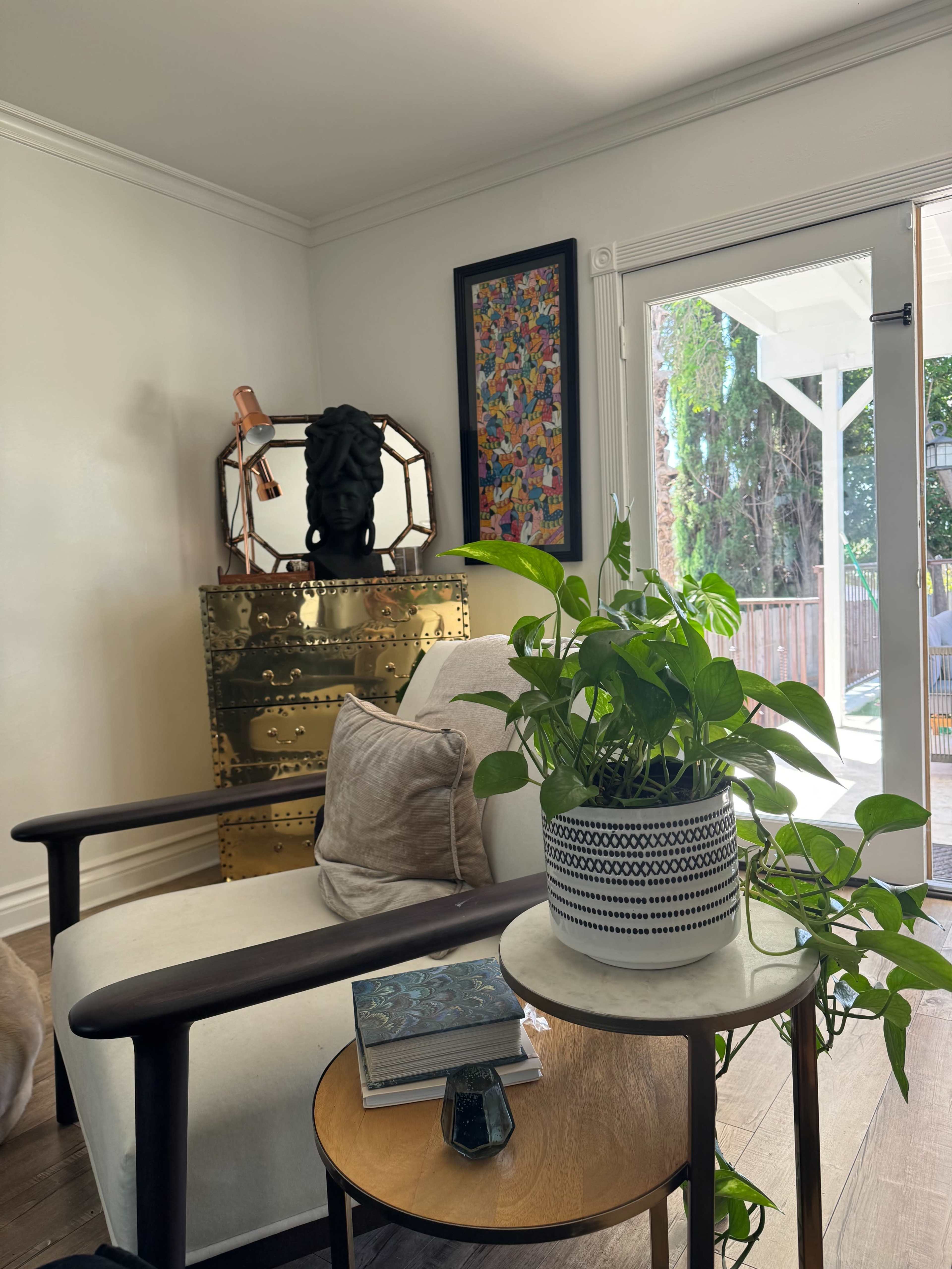 A cozy living room features a chair beside a small table with a plant, books, and decorative items, along with a gold accent cabinet and artwork on the wall.