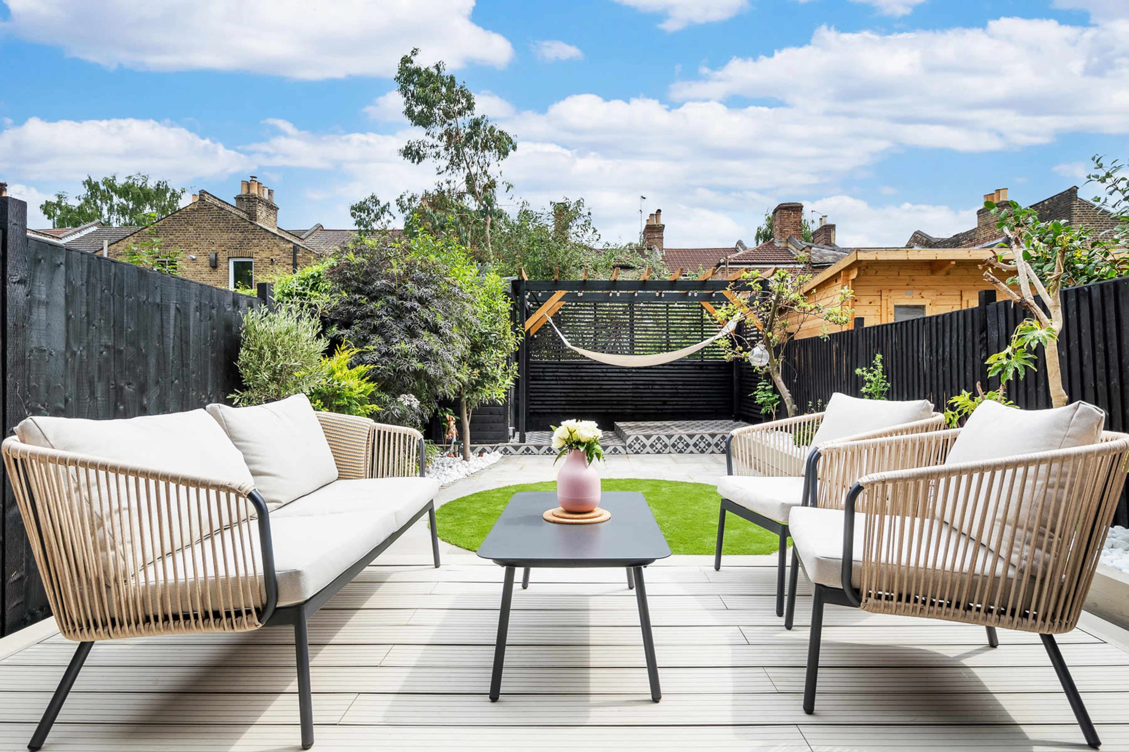 A landscaped garden area with two modern seating arrangements and a small table, featuring a hammock in the background and greenery along the perimeter.
