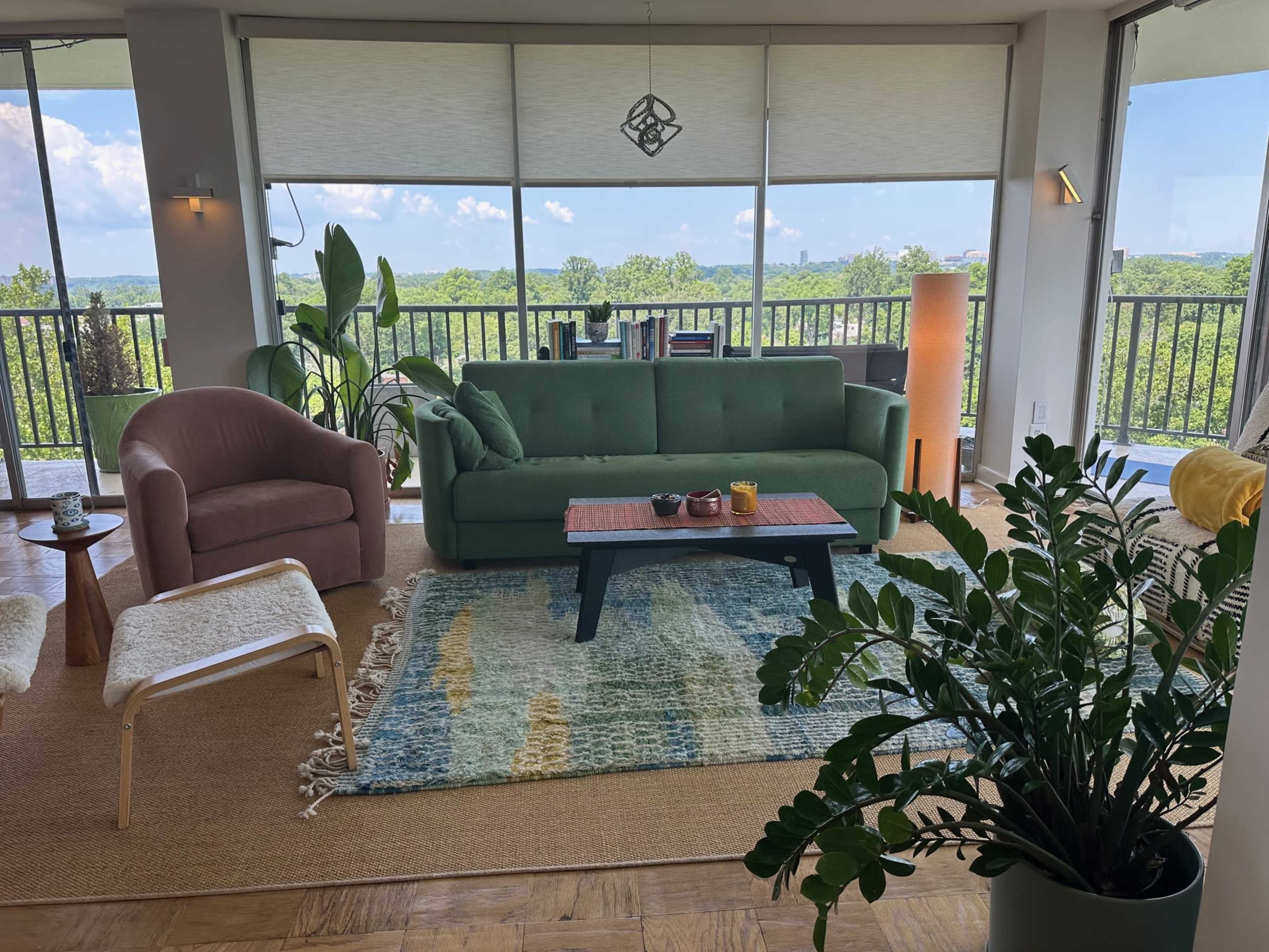 The image shows a modern living room with a green sofa, a pink armchair, a coffee table, and a view of greenery outside through large windows.