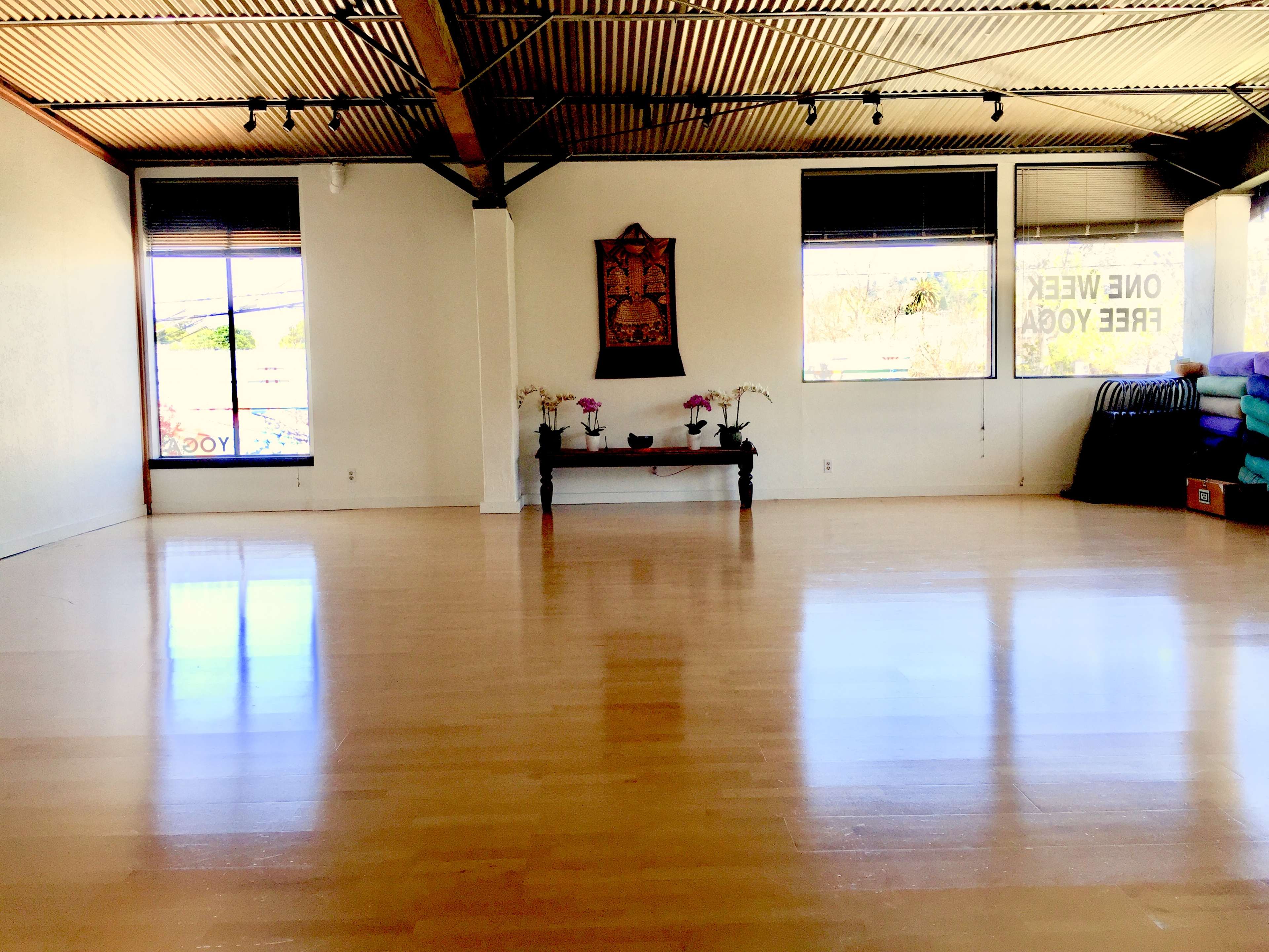 An empty, well-lit yoga studio with polished wooden floors and a decorative wall hanging above a table with orchids.