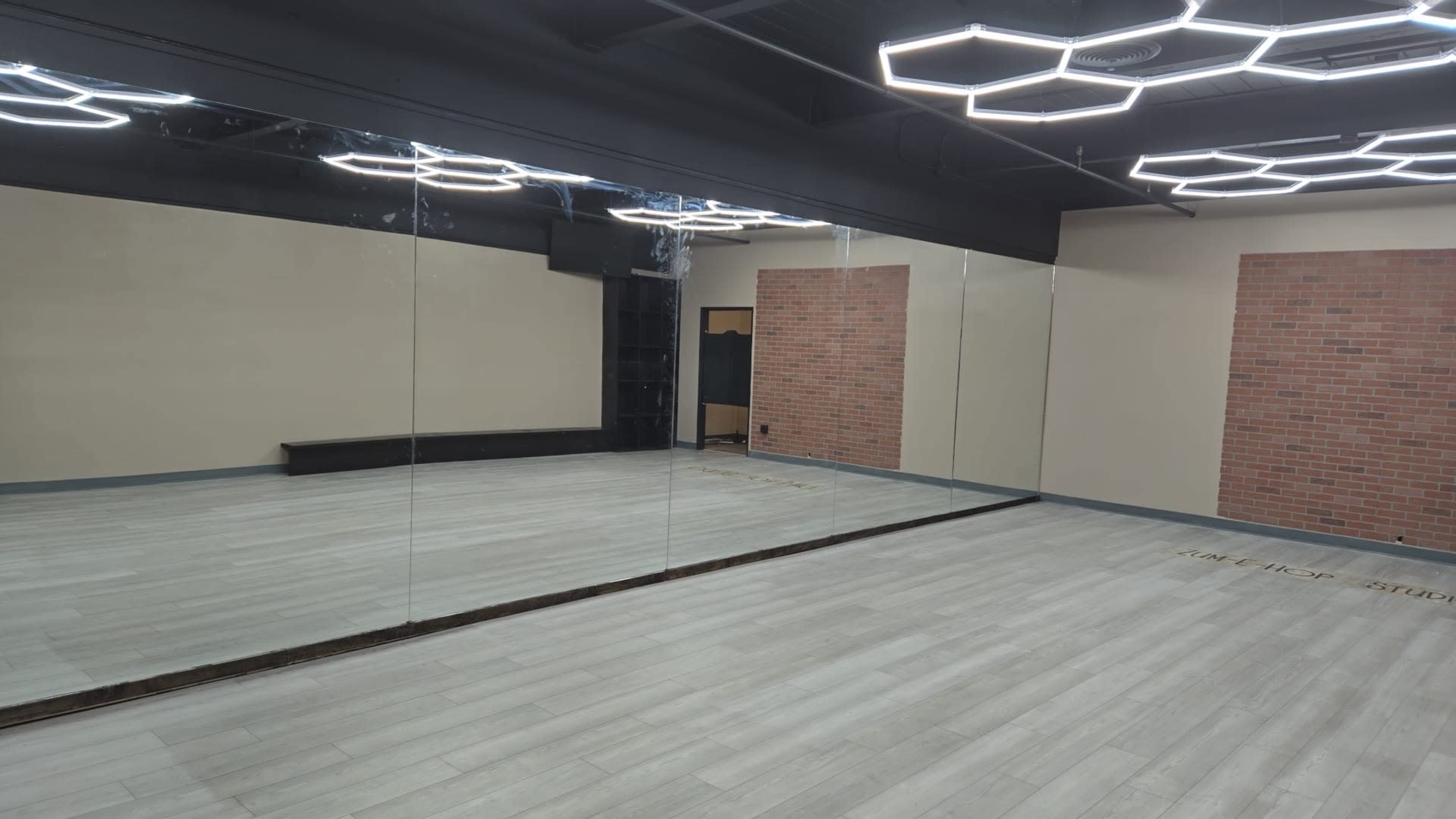 The image shows a spacious room with light-colored flooring, large mirrors on one wall, and modern lighting fixtures hanging from the ceiling.