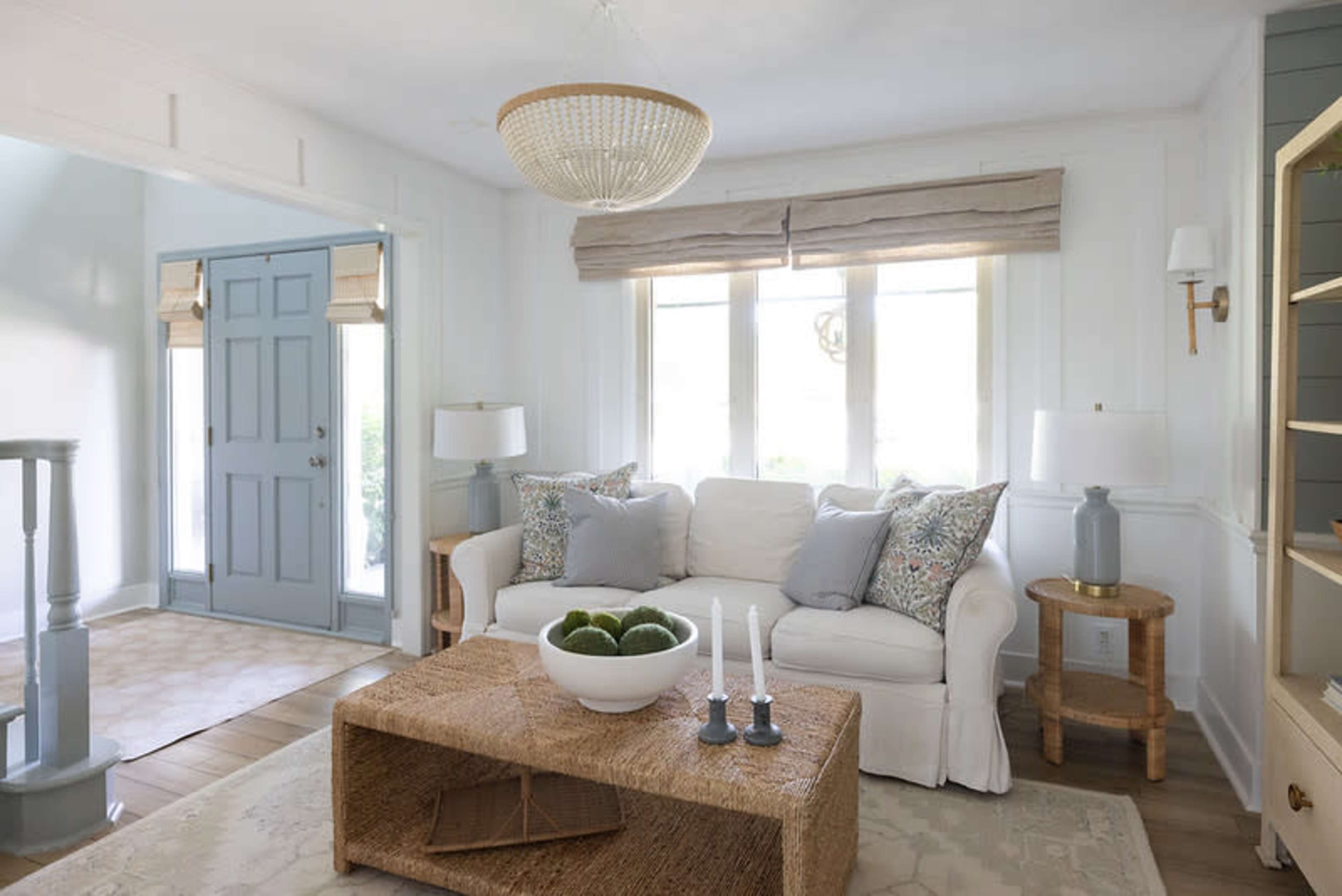 The image shows a bright living room featuring a white sofa with decorative pillows, a natural fiber coffee table, and large windows adorned with light-colored shades.