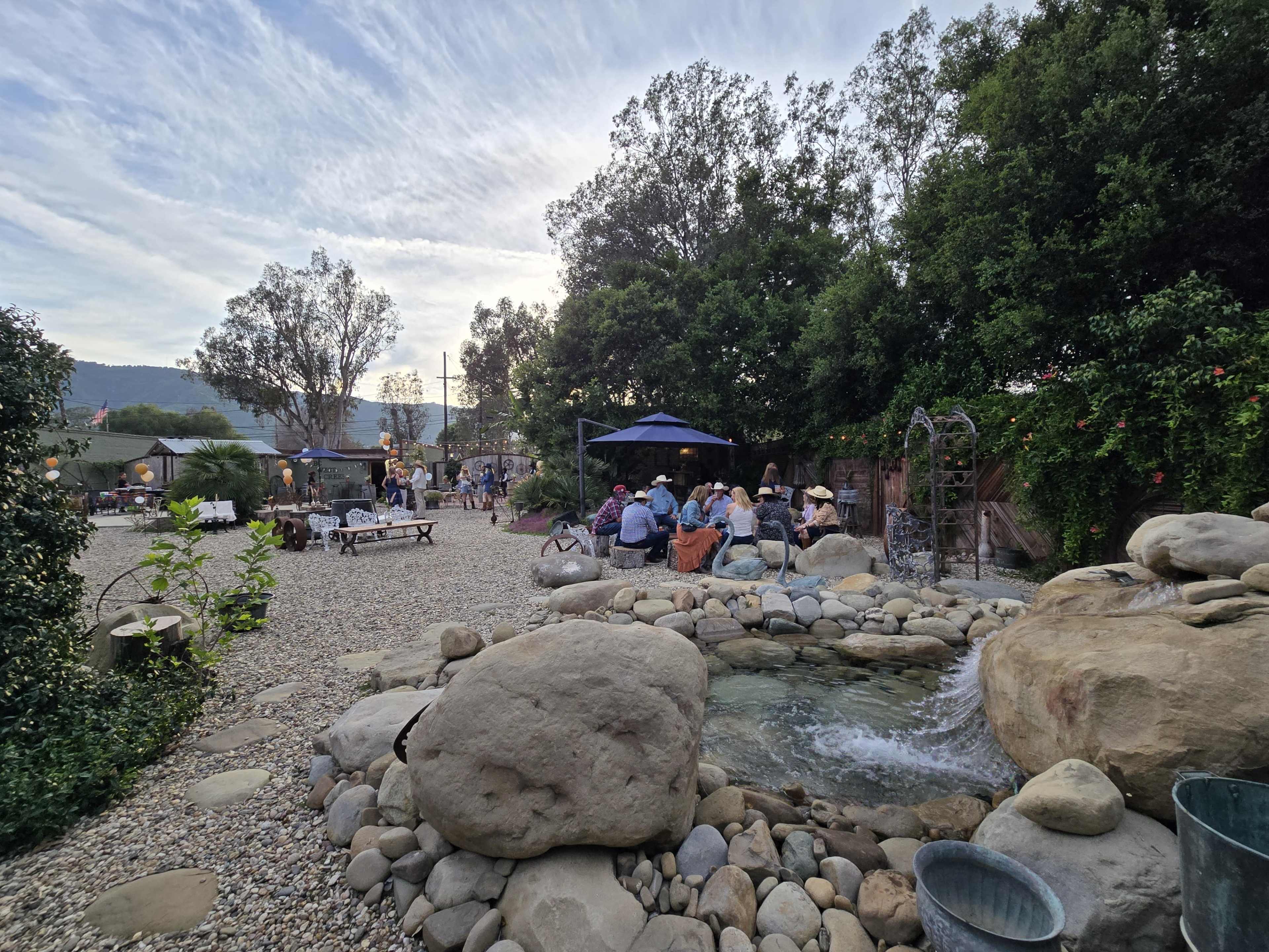 A gathering space with people seated around tables, a small water feature, and greenery, surrounded by gravel paths and trees.
