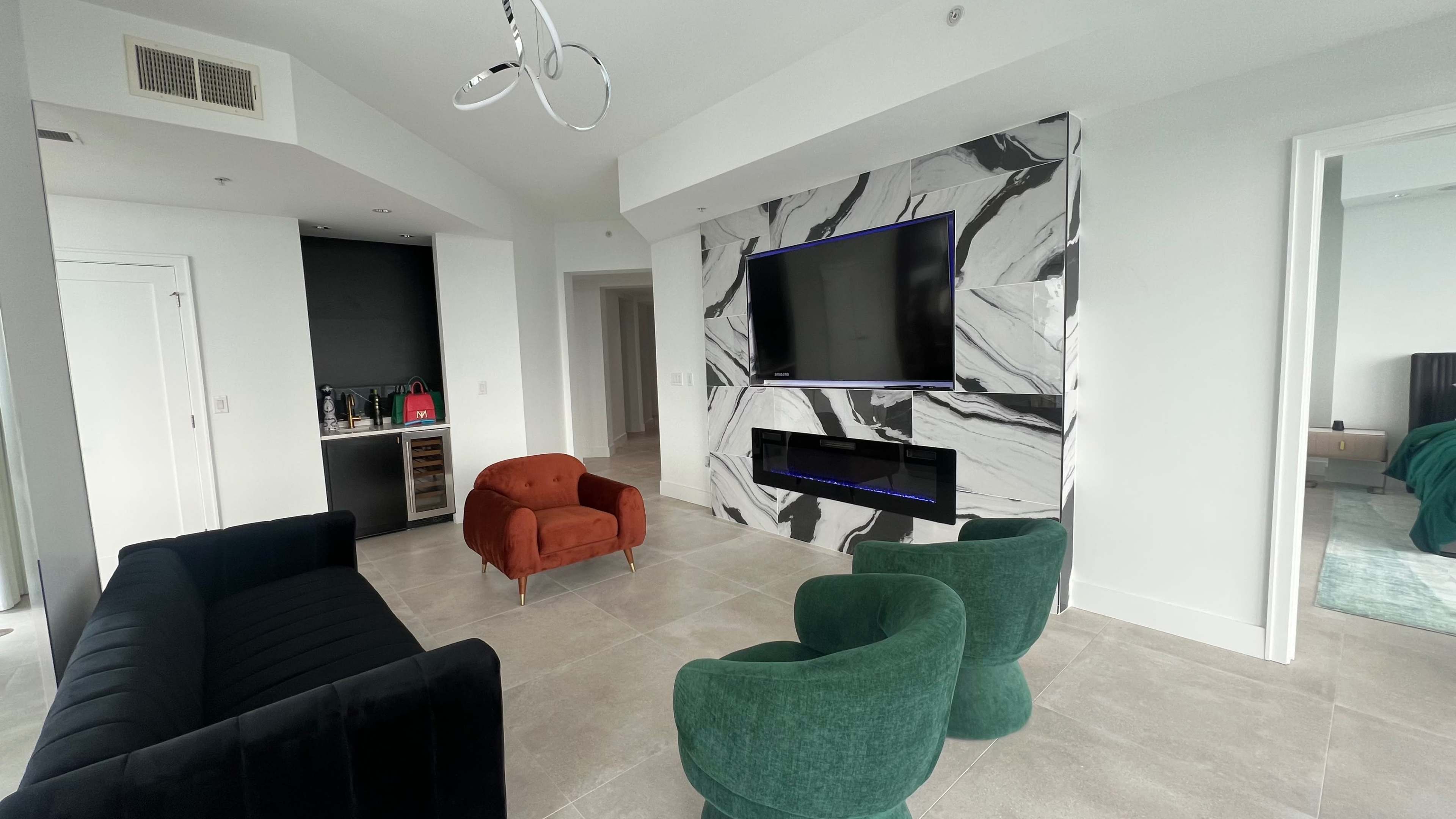 The image shows a modern living space with a marble-featured wall, a wall-mounted television, two green chairs, an orange armchair, and a black sofa.