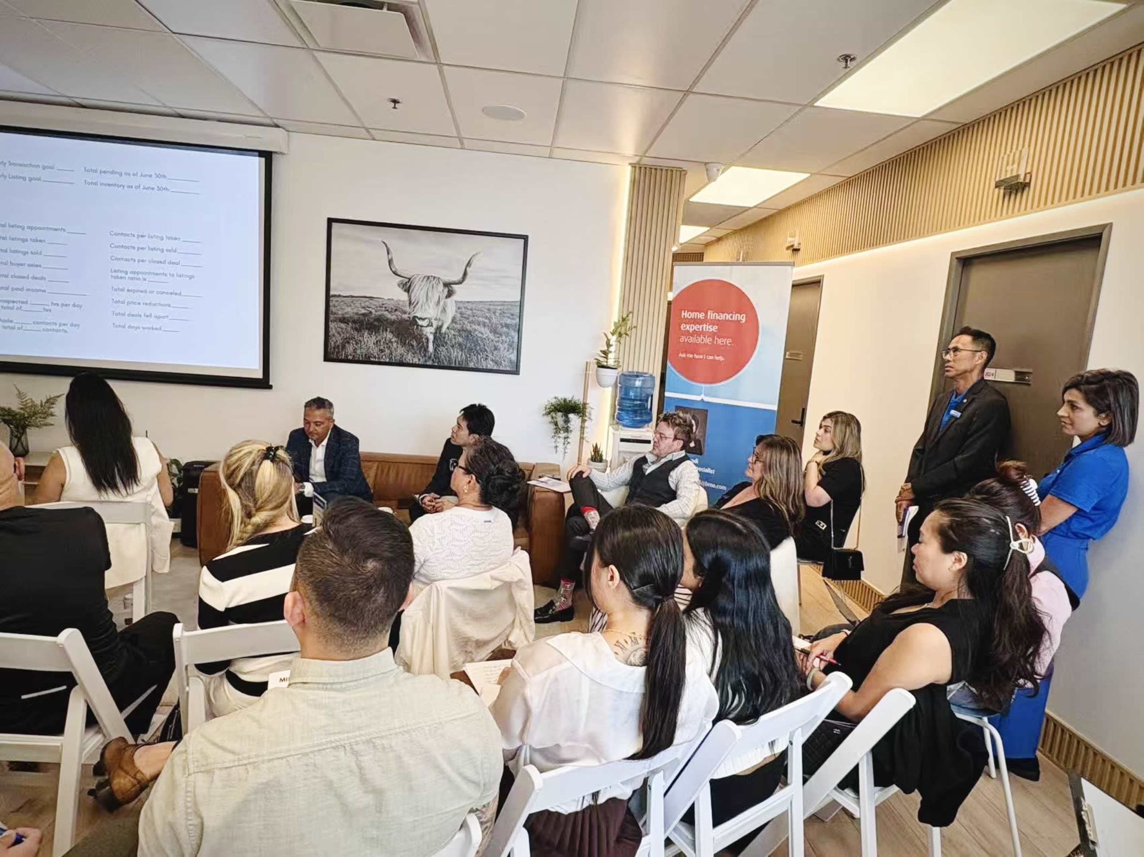 A group of people is seated and standing in a conference room, attentively listening to a presentation displayed on a screen.