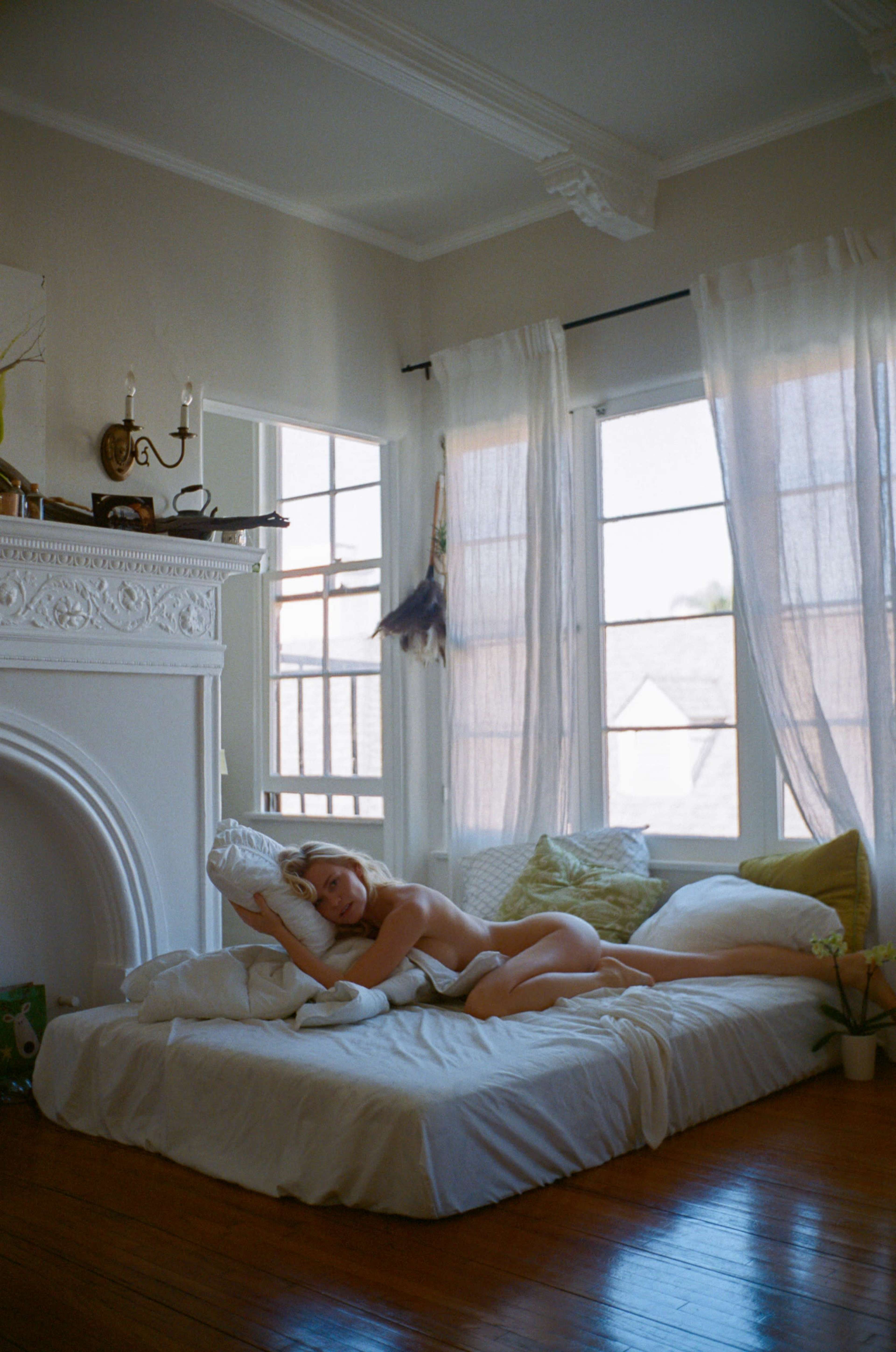 A person is lying on a bed in a well-lit room with large windows and light-colored curtains.
