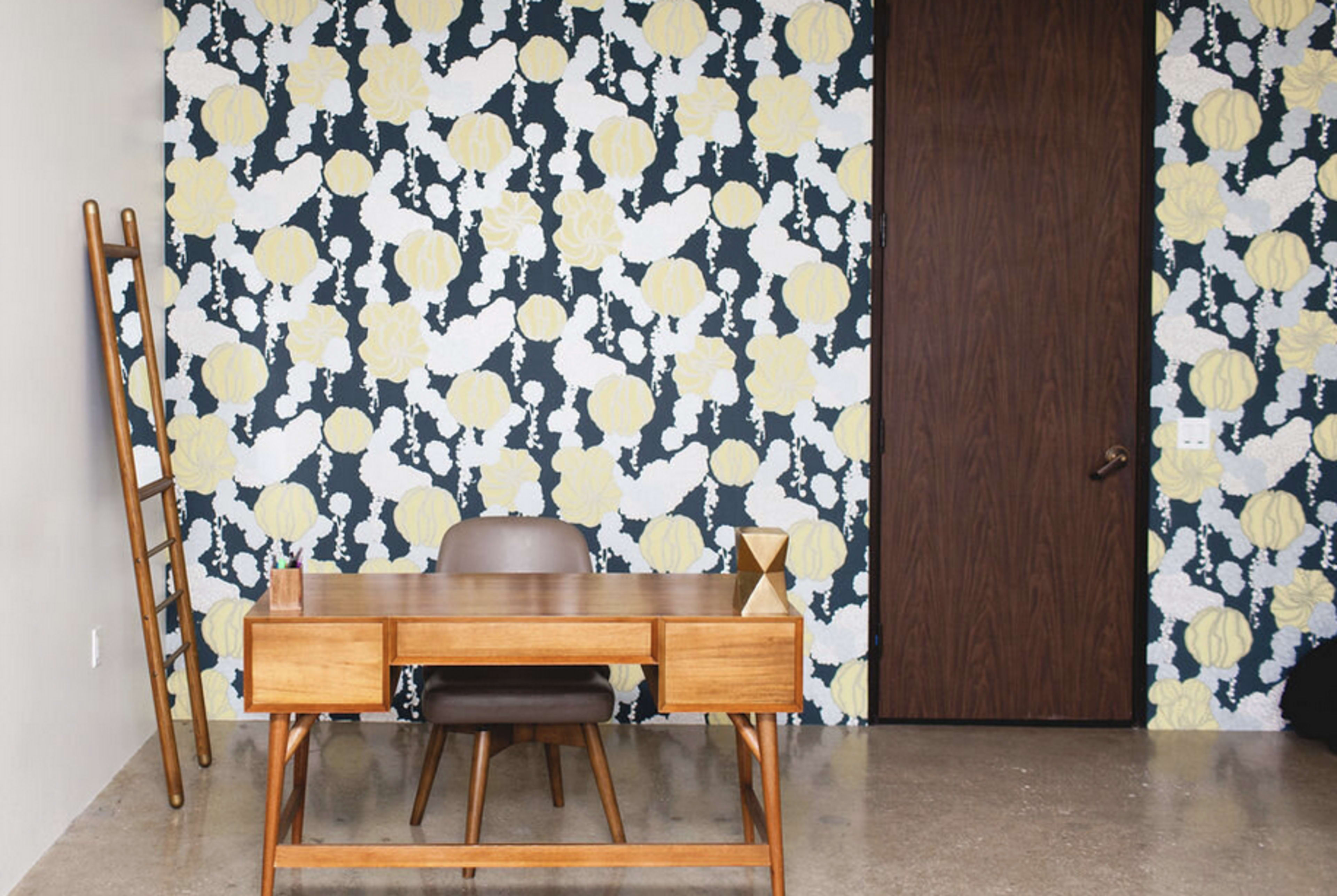 A workspace featuring a wooden desk and chair against a floral-patterned wallpaper and a closed door.