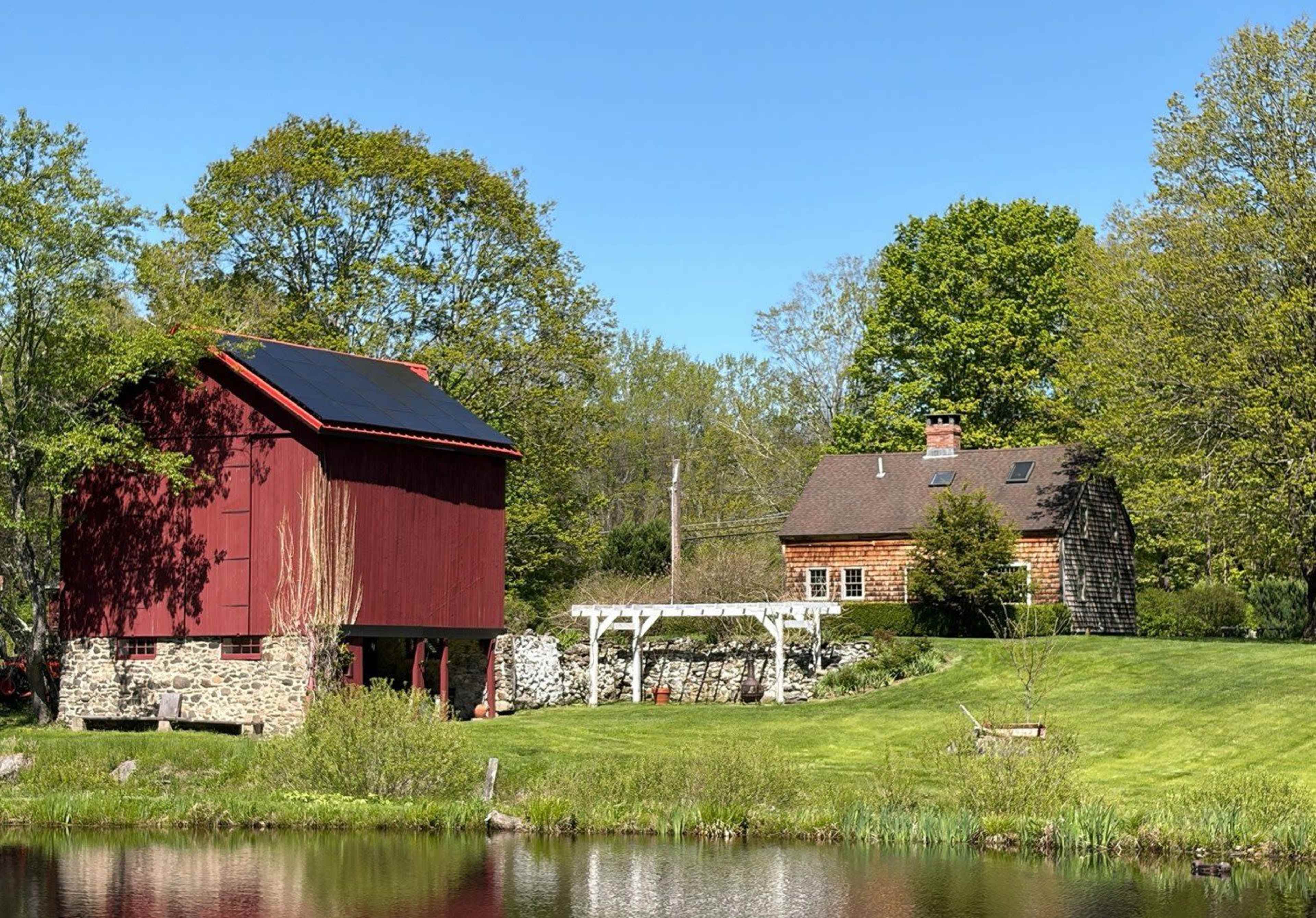 A red barn with solar panels stands next to a quaint wooden house by a pond, surrounded by lush greenery.