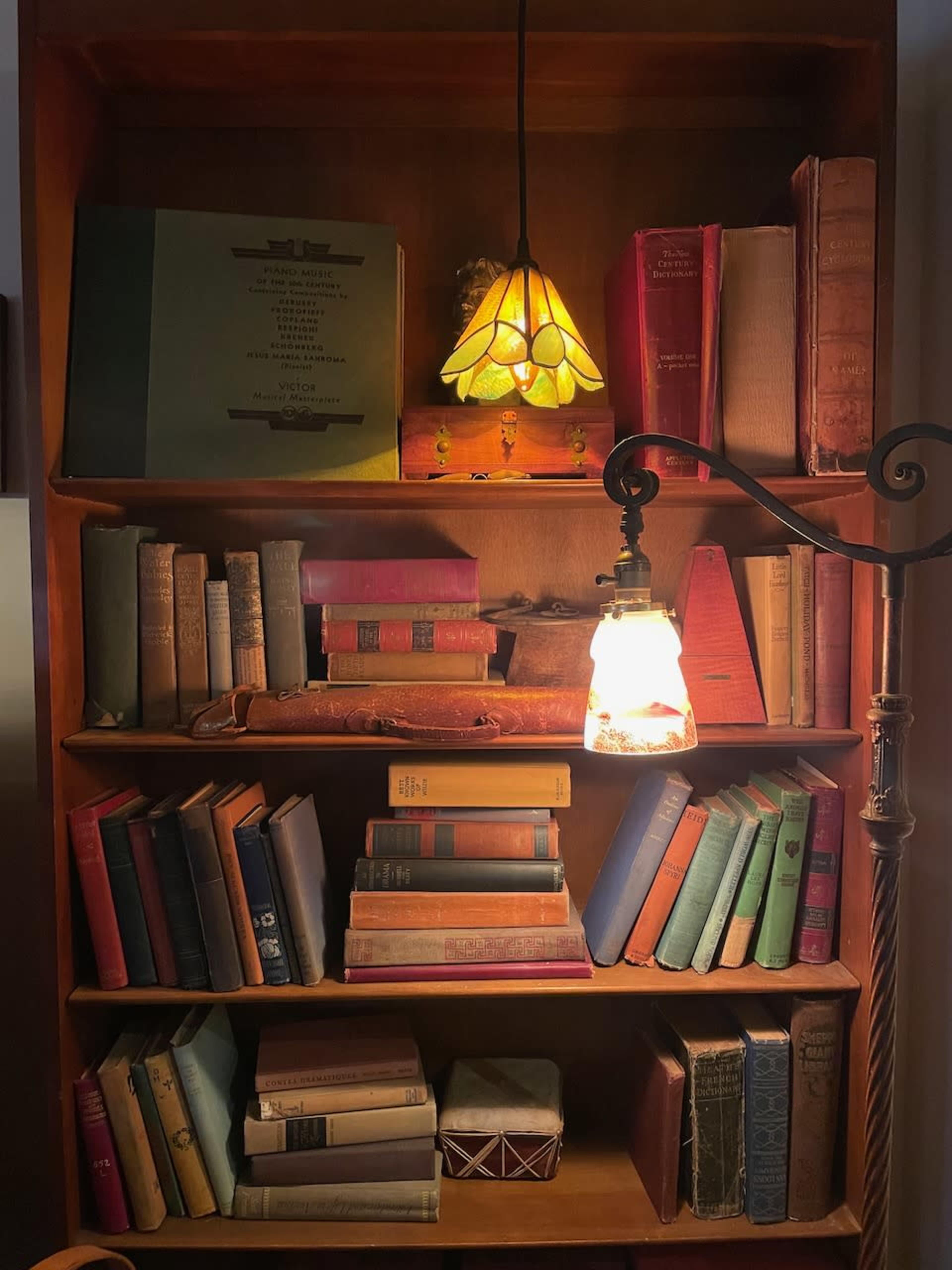 A wooden bookshelf displays an array of old books, illuminated by a stained glass lamp and a hanging light.