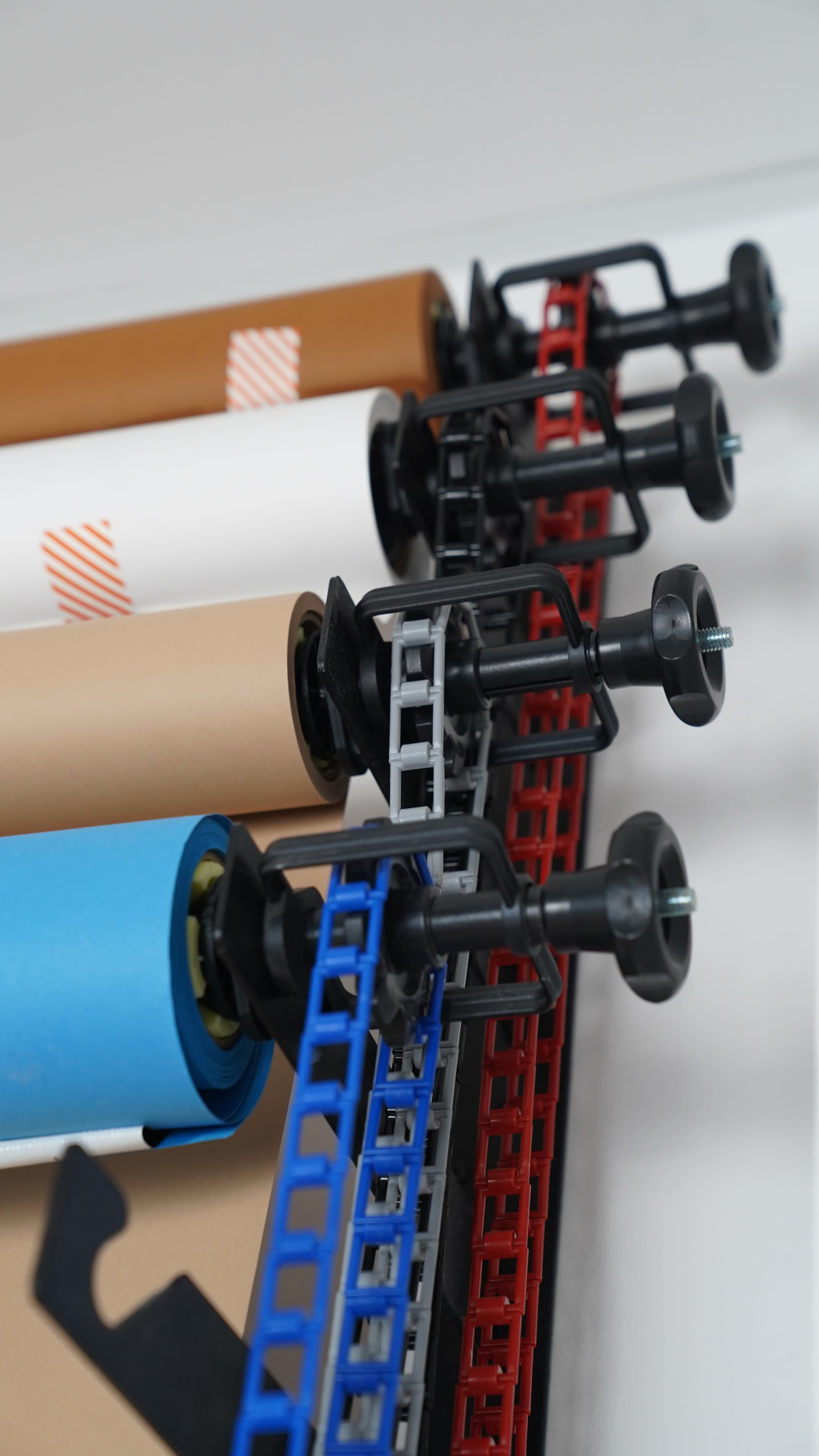 The image shows a row of paper rolls in various colors mounted on a wall rack with adjustable holders.