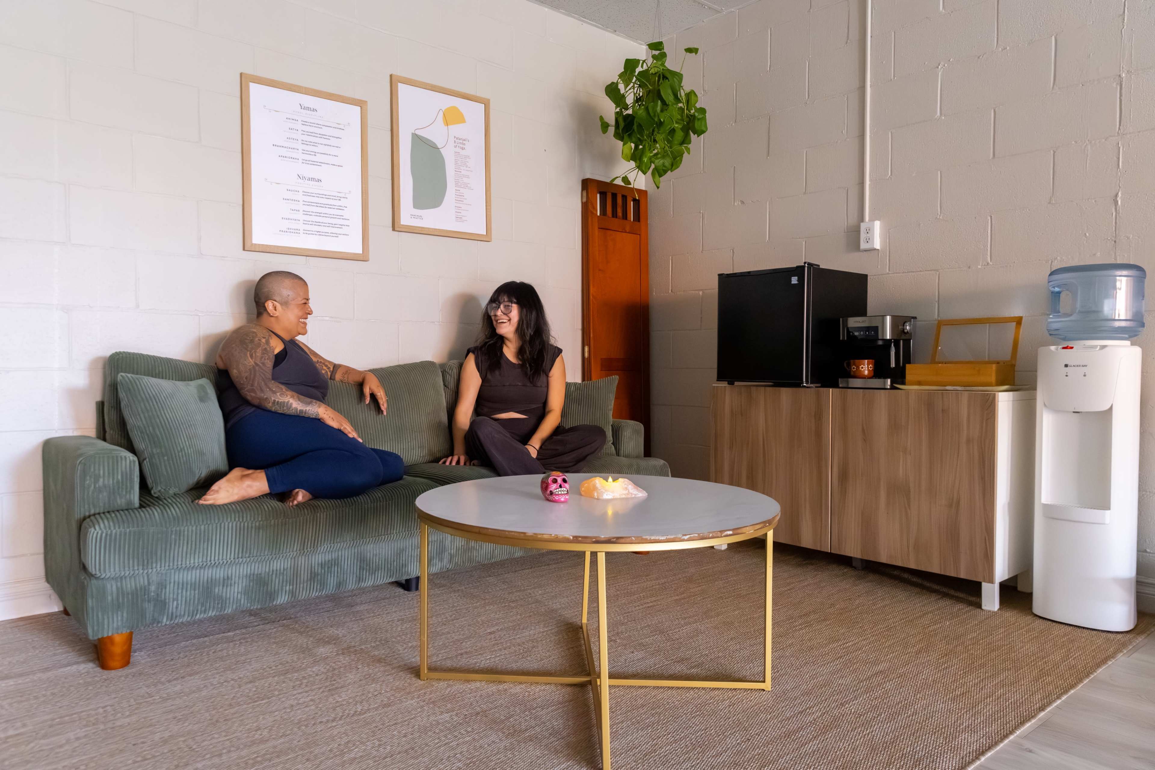 Two women are sitting on a green couch in a modern, light-colored living space that includes a coffee maker, water cooler, and decorative plants.