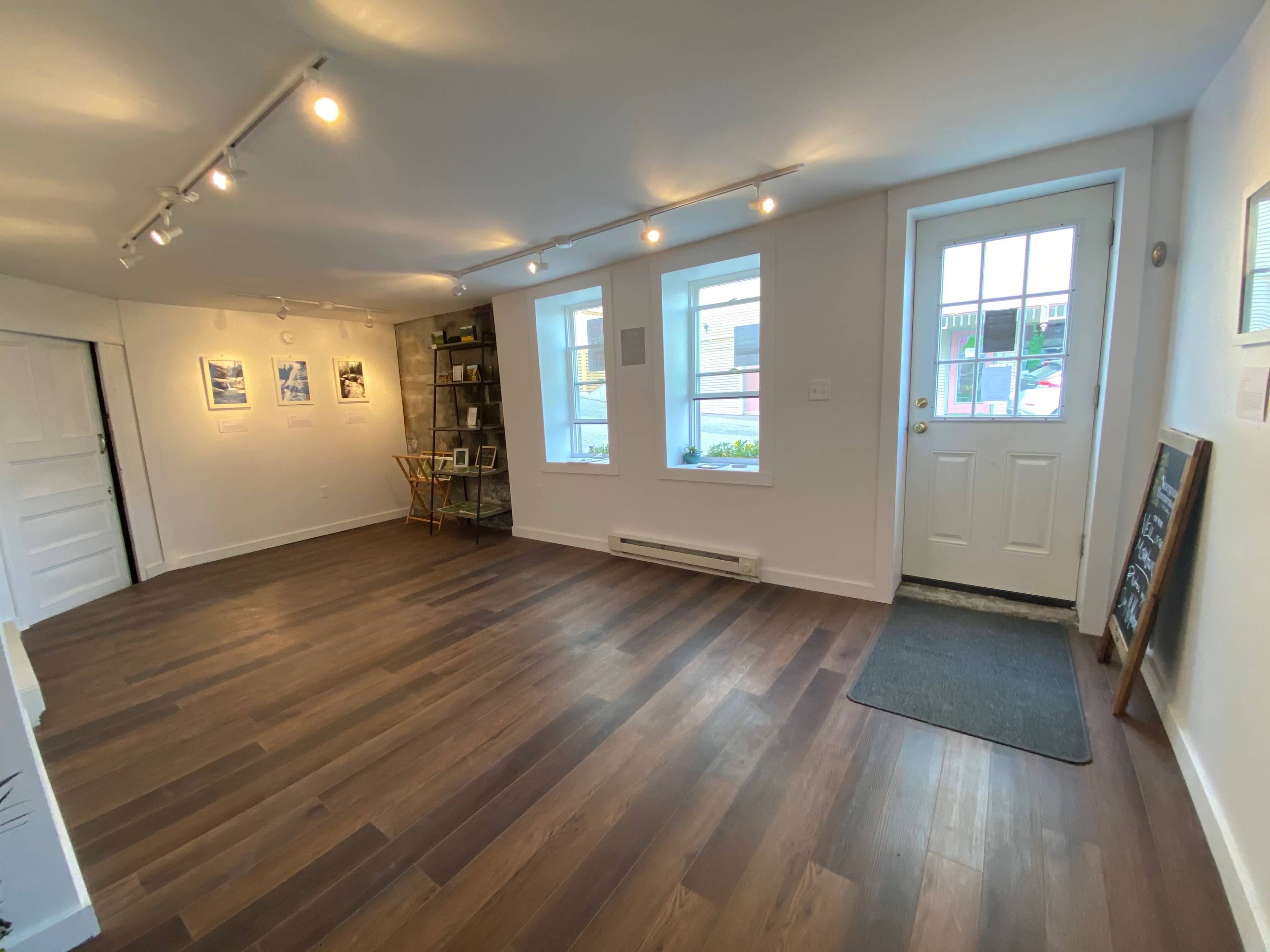 A bright, minimally furnished room with wooden flooring, three windows, and a door leading outside.
