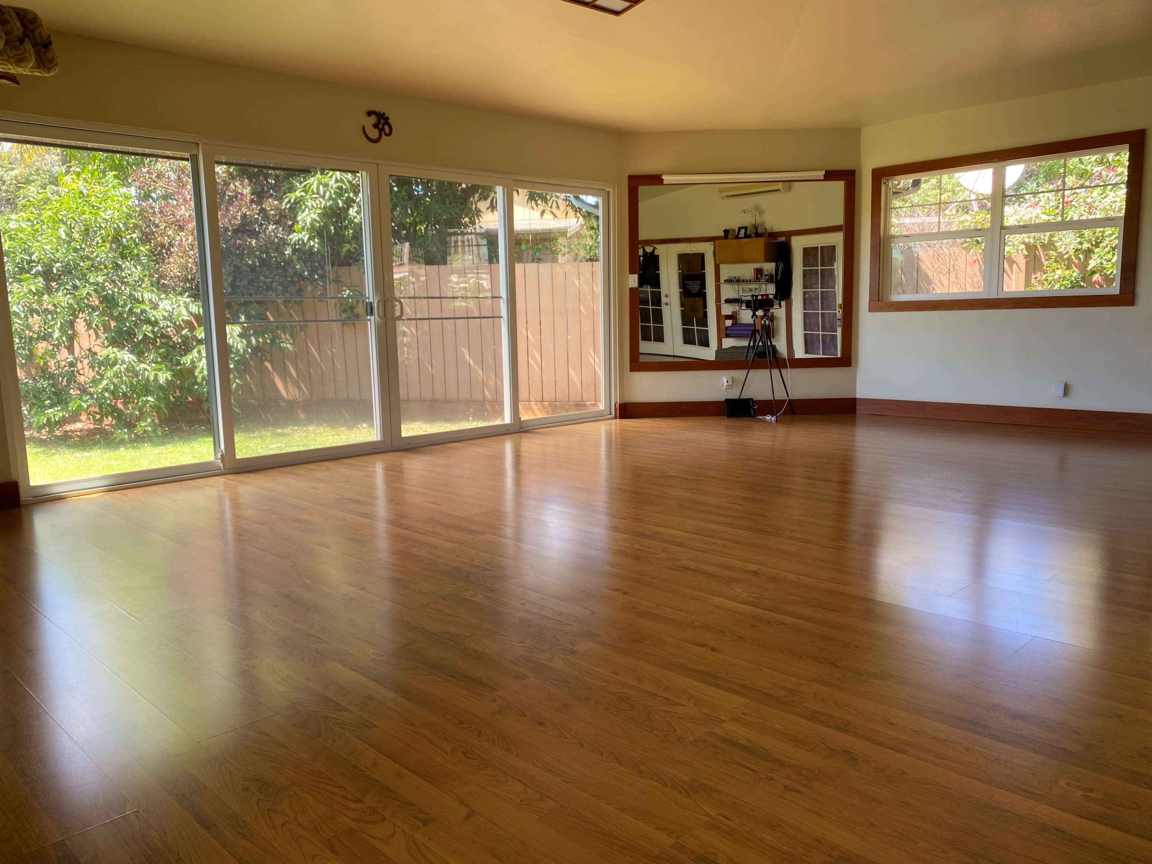 A spacious, sunlit room with wooden flooring, large windows, and a mirror reflecting the outdoor greenery.