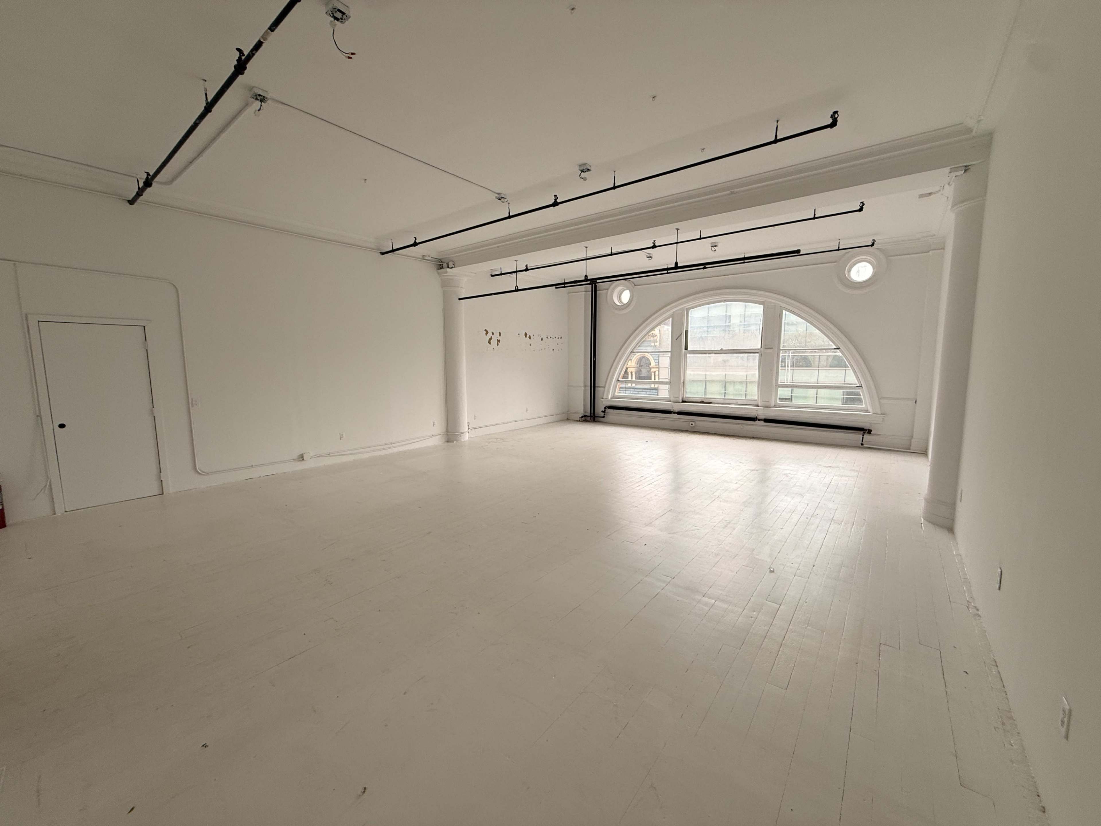 The image shows a large, empty room with white walls and a wooden floor, featuring a large arched window letting in natural light.