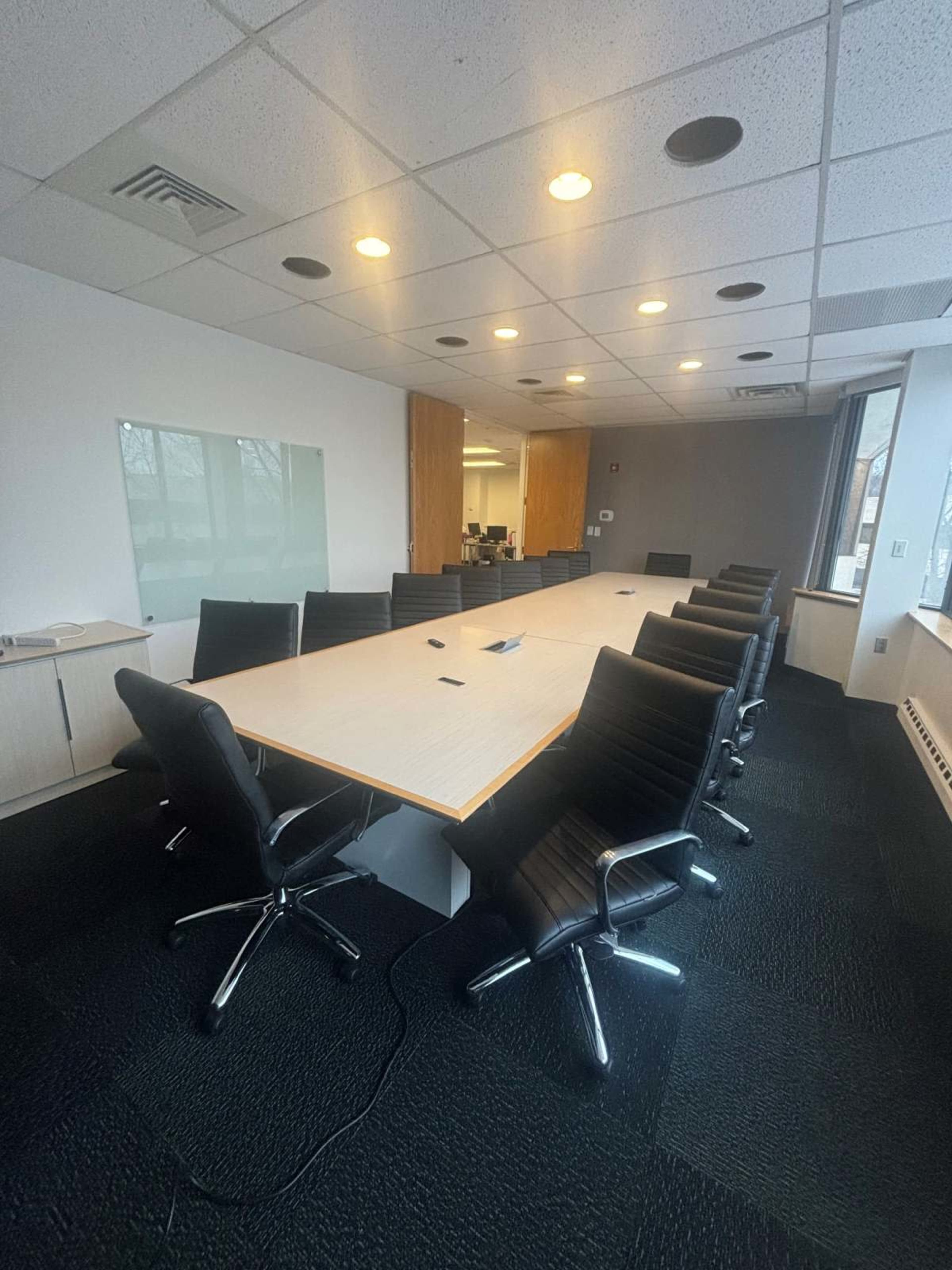 The image shows a large conference room with a long table surrounded by black office chairs, illuminated by overhead lights.