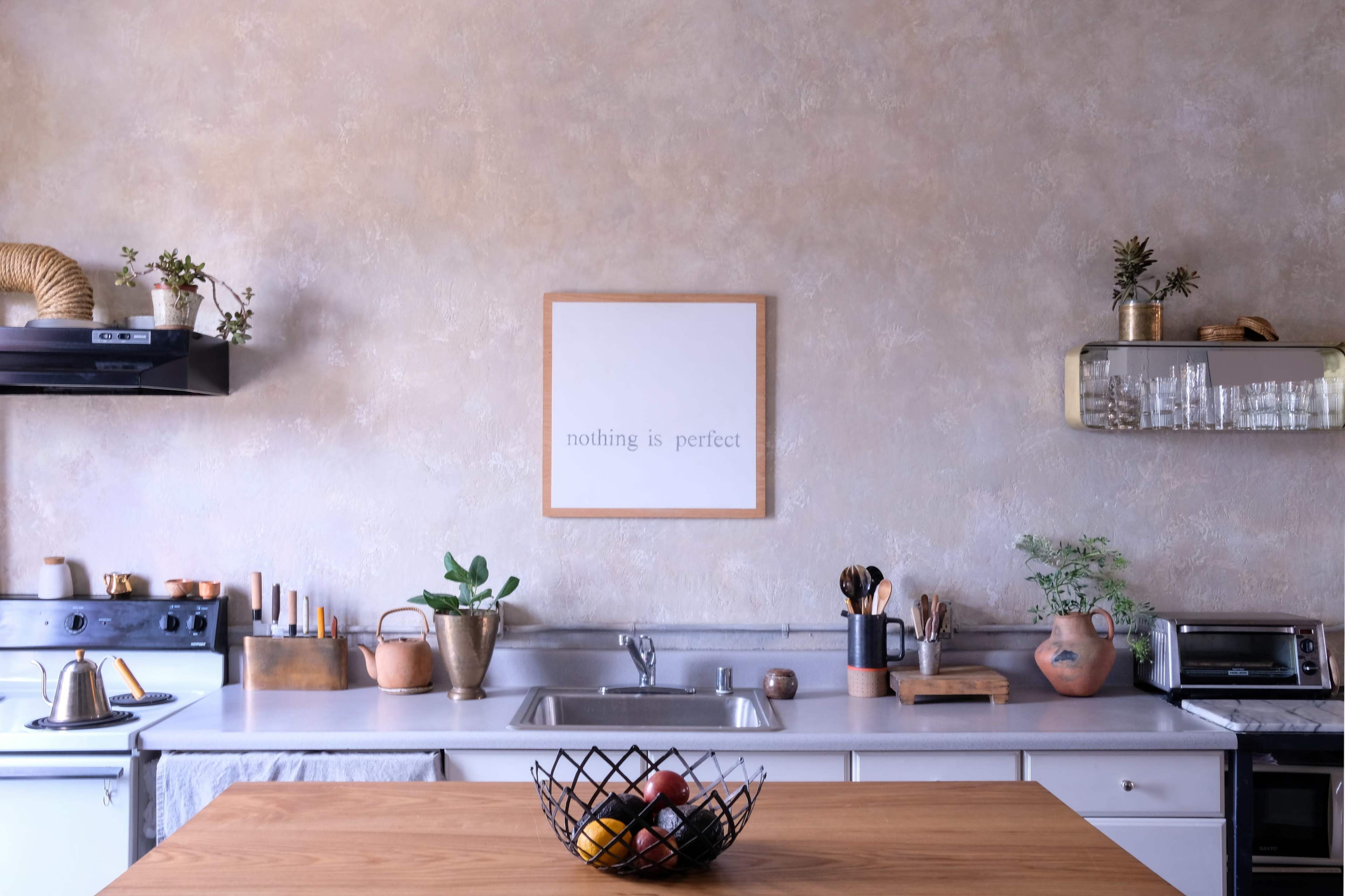 A modern kitchen features a wooden table, a sink with utensils, and a decorative wall with a framed quote.